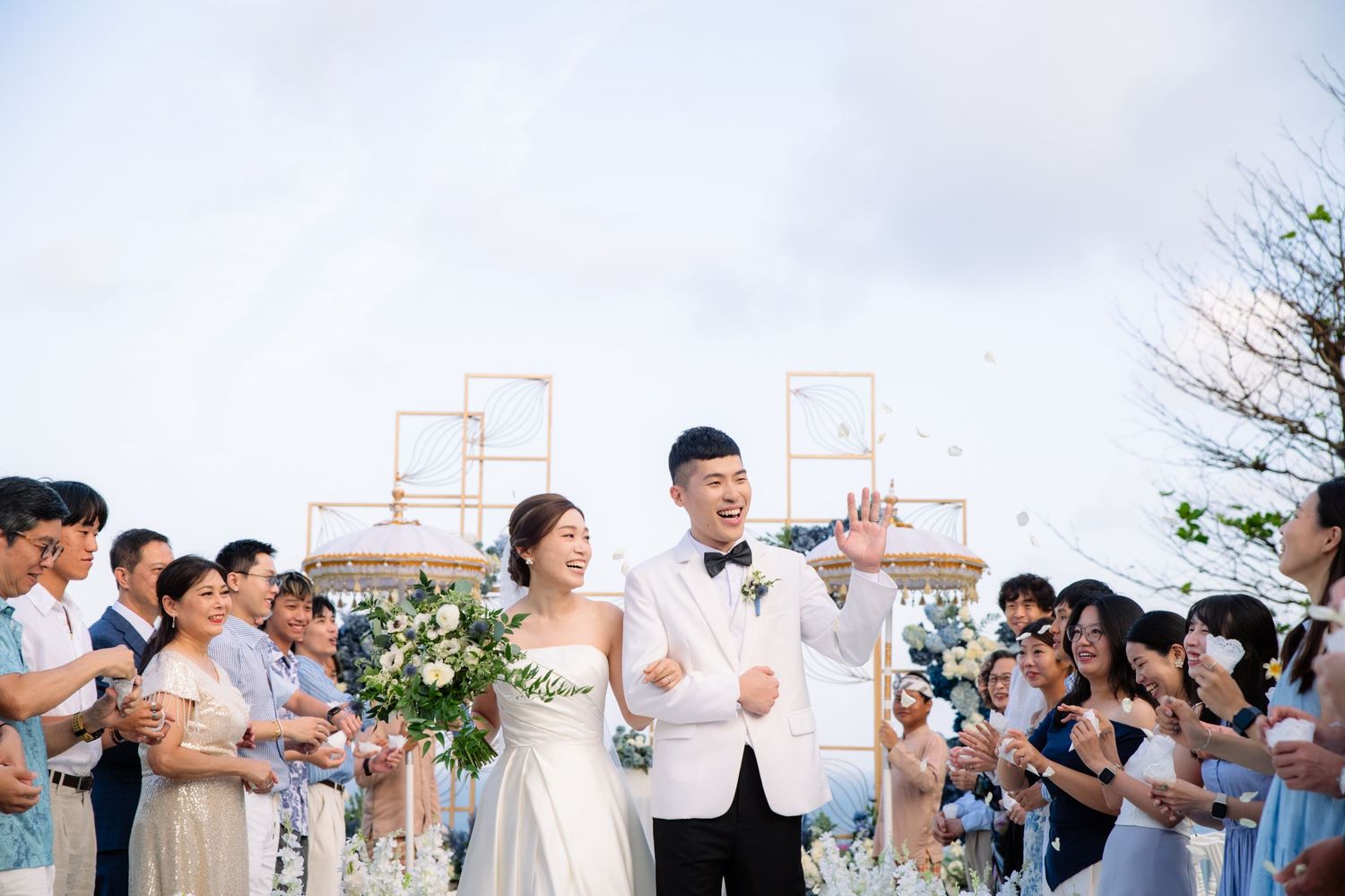 Bride in a white gown holding a bouquet walks with a groom in a white tuxedo past cheering wedding guests at an outdoor ceremony.