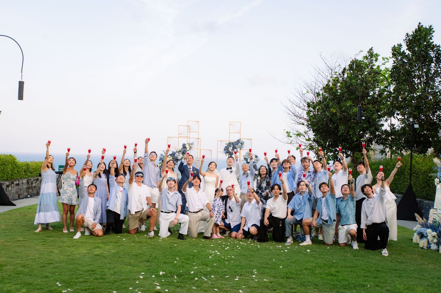 Large wedding group in light blue and white attire cheers with raised arms on a green lawn at an oceanside venue.