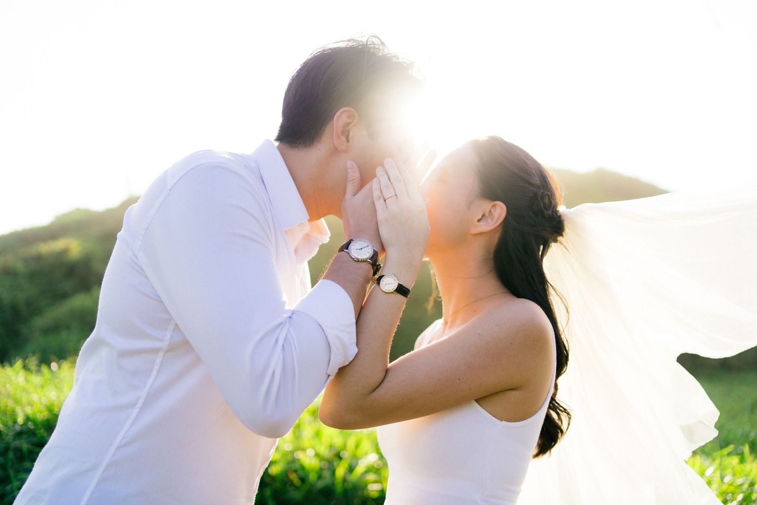 Couple in white wedding attire sharing a romantic moment outdoors with sunlight glowing behind them.