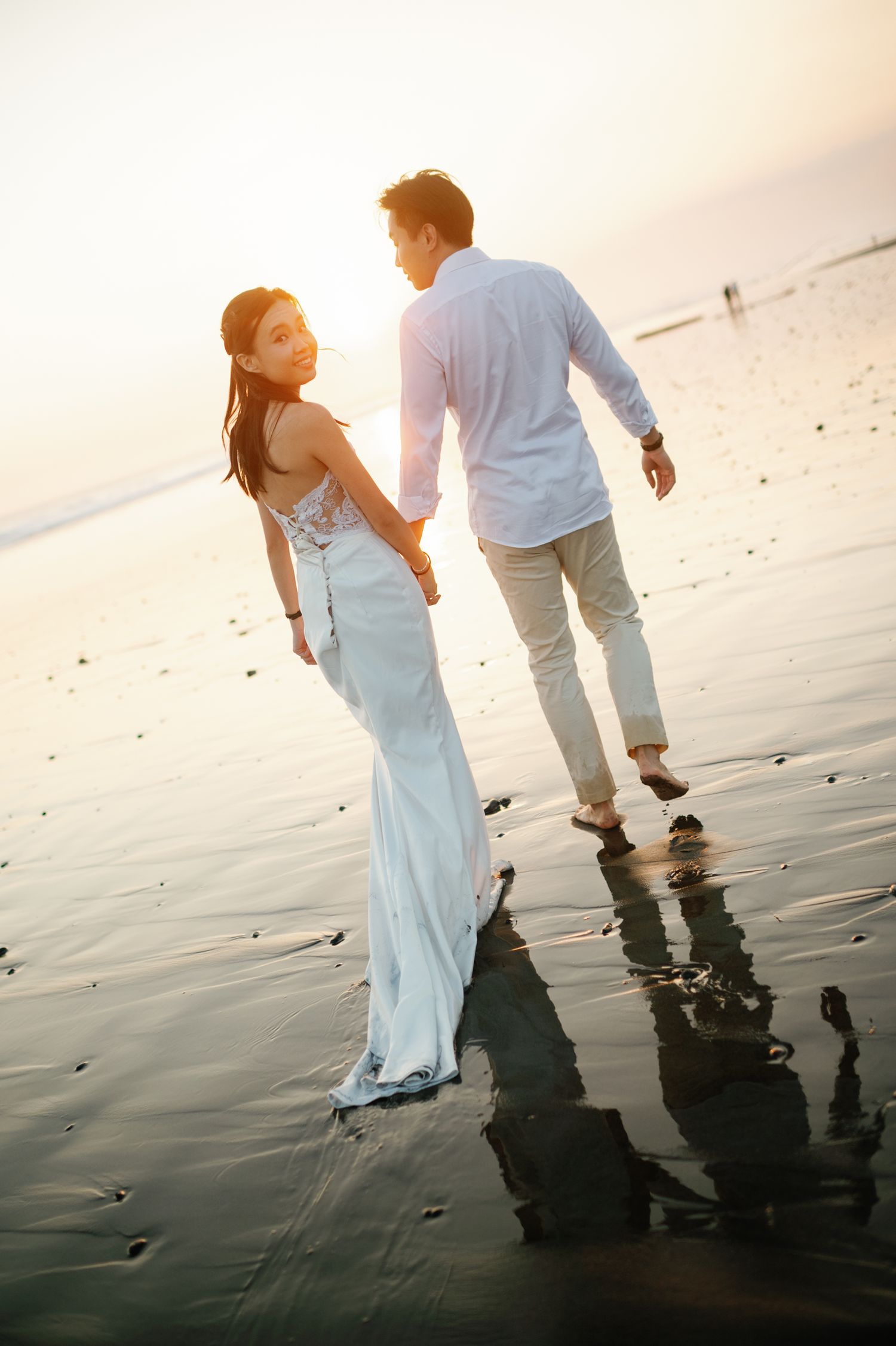 Couple holding hands on a reflective beach at sunset, woman in white gown looking back.