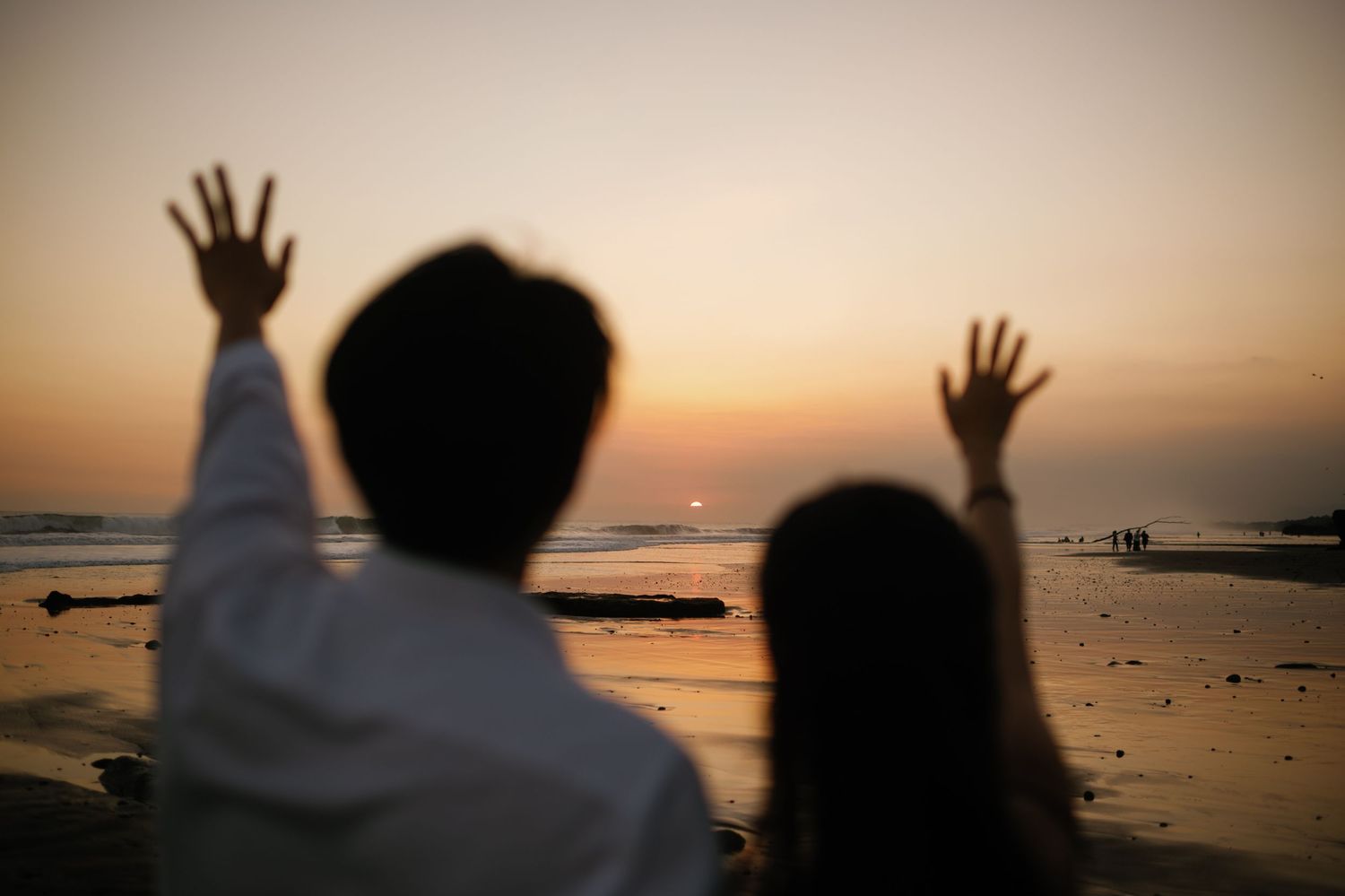 Couple raising hands toward a golden sunset over the ocean beach, viewed from behind in silhouette.