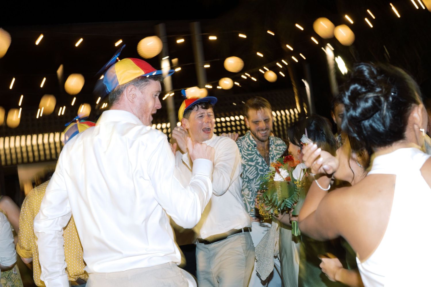 Guests dancing at a festive outdoor party with colorful hats and string lights in the background.