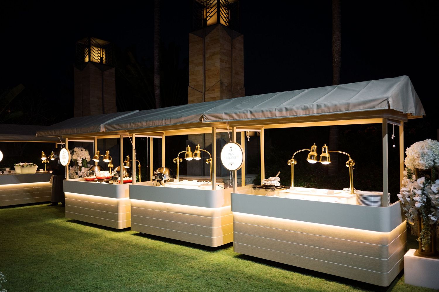 Elegant outdoor event booth with gold accents and pendant lights set up on green grass at night.