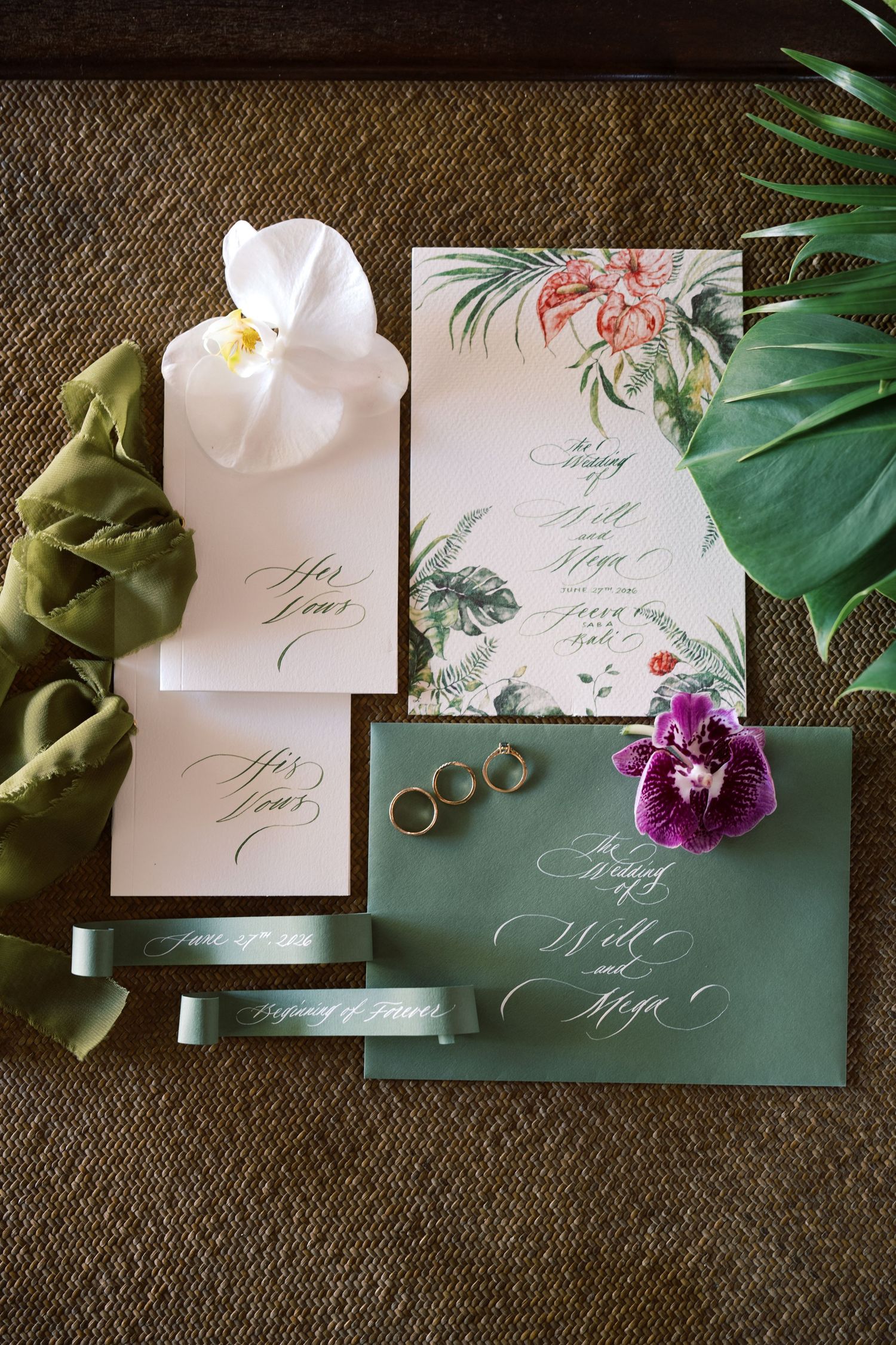 Tropical wedding invitation suite with green envelopes, floral cards, gold rings, and orchids on a woven surface.