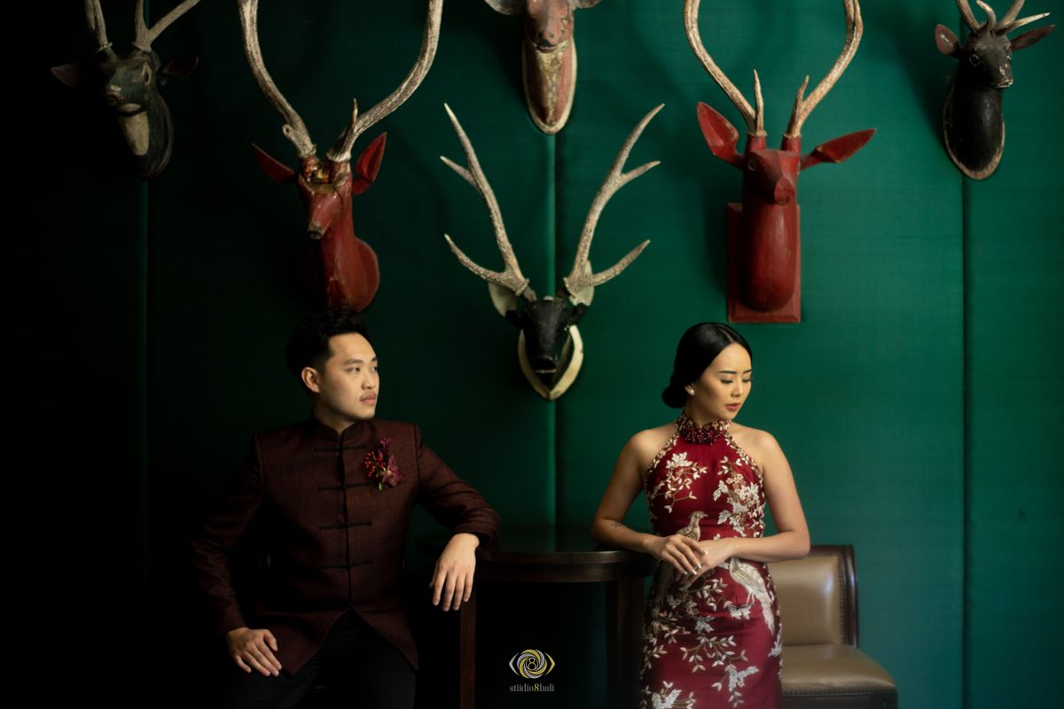 Couple in red traditional Chinese attire seated against green wall adorned with decorative deer antler mounts.