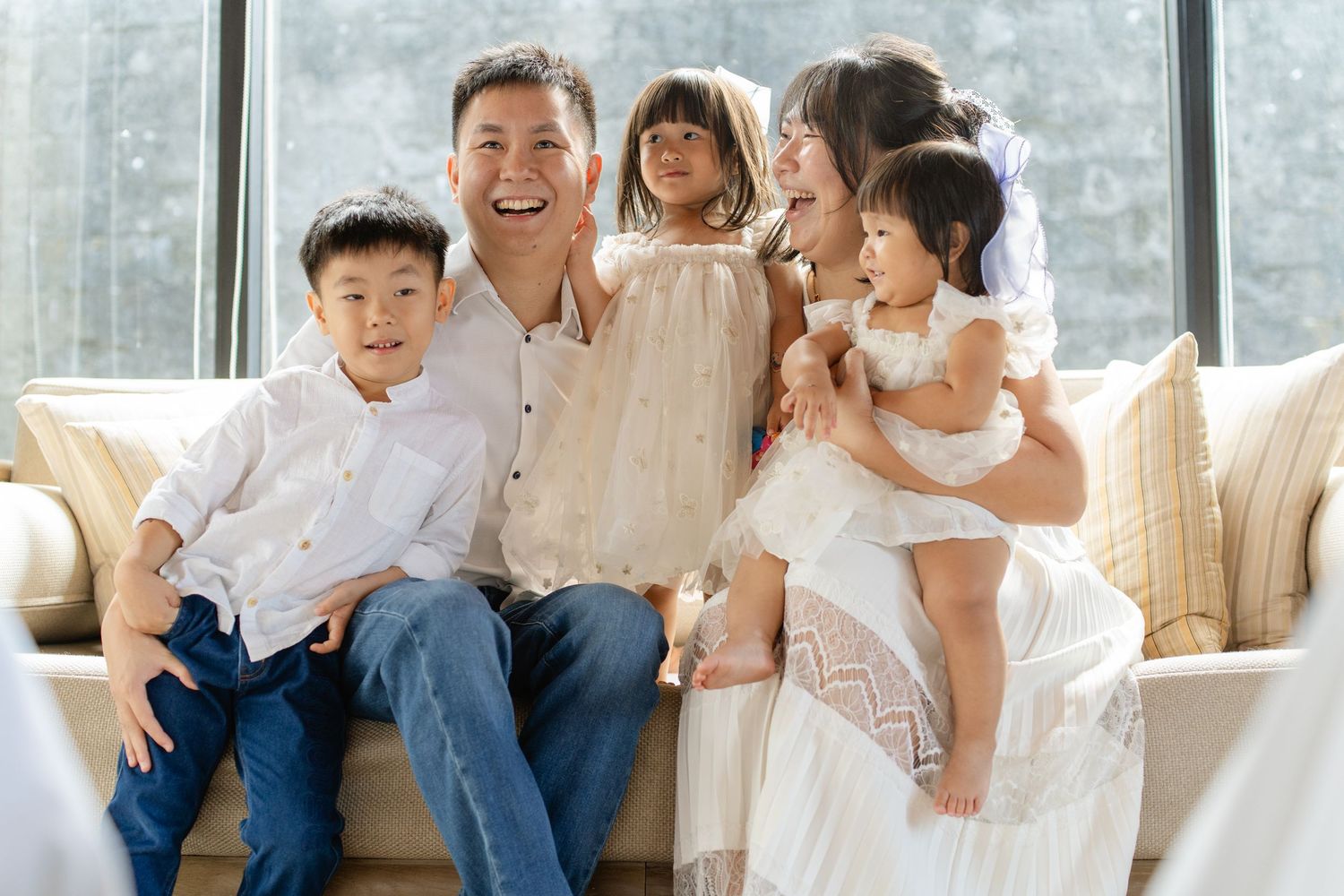 Happy Asian family with three young children smiling together on a couch in a bright, elegant living room.