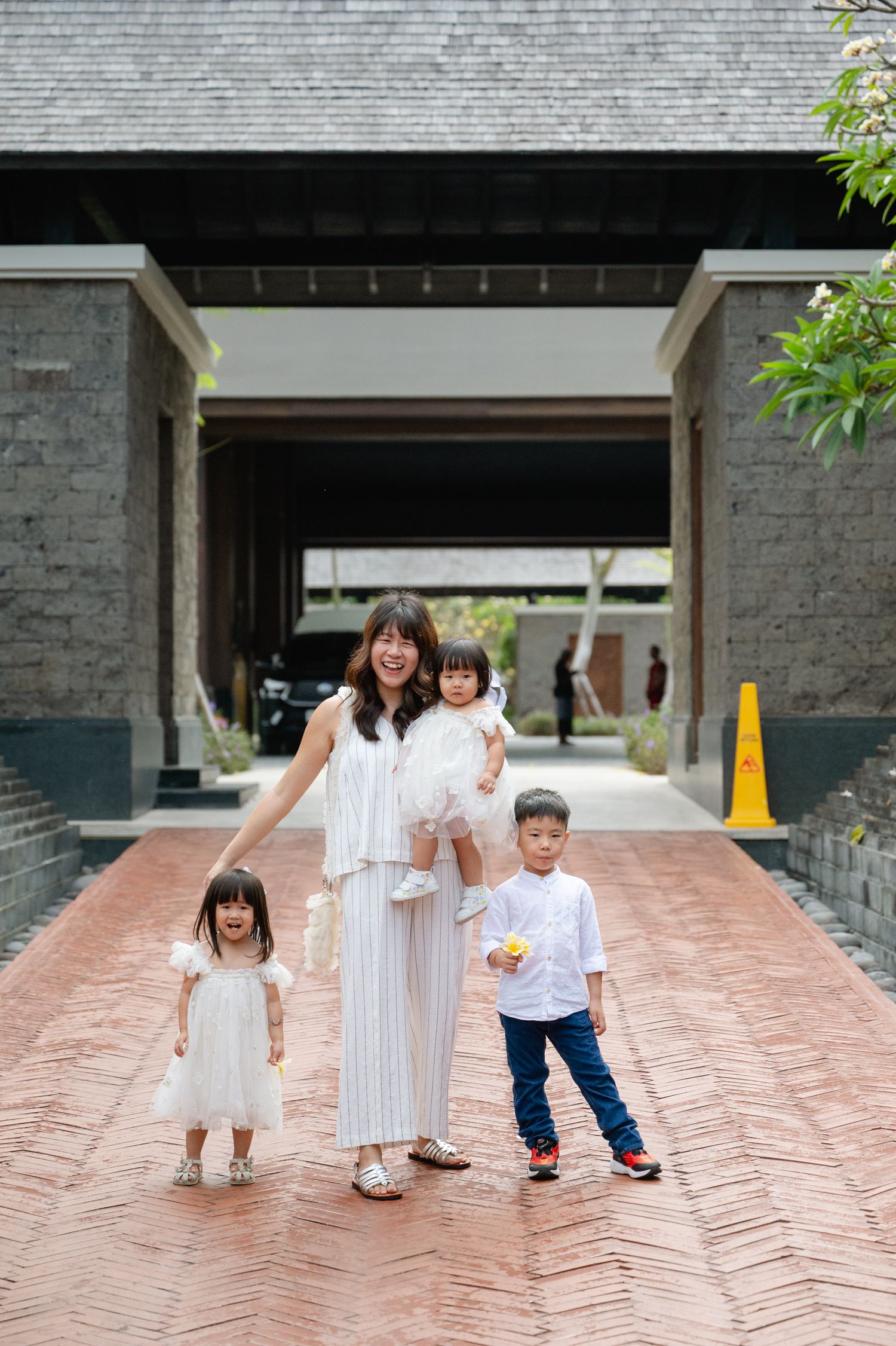 A smiling woman in white holds a toddler while two young children stand beside her on a brick path at a resort.