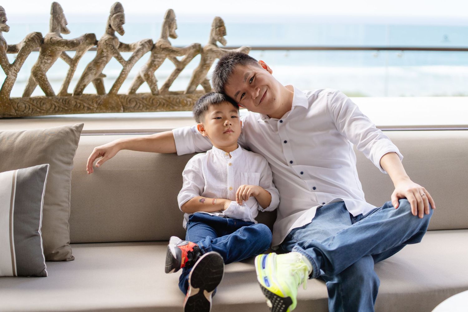 A man and young boy smile together on a couch with ocean views in a modern living space.