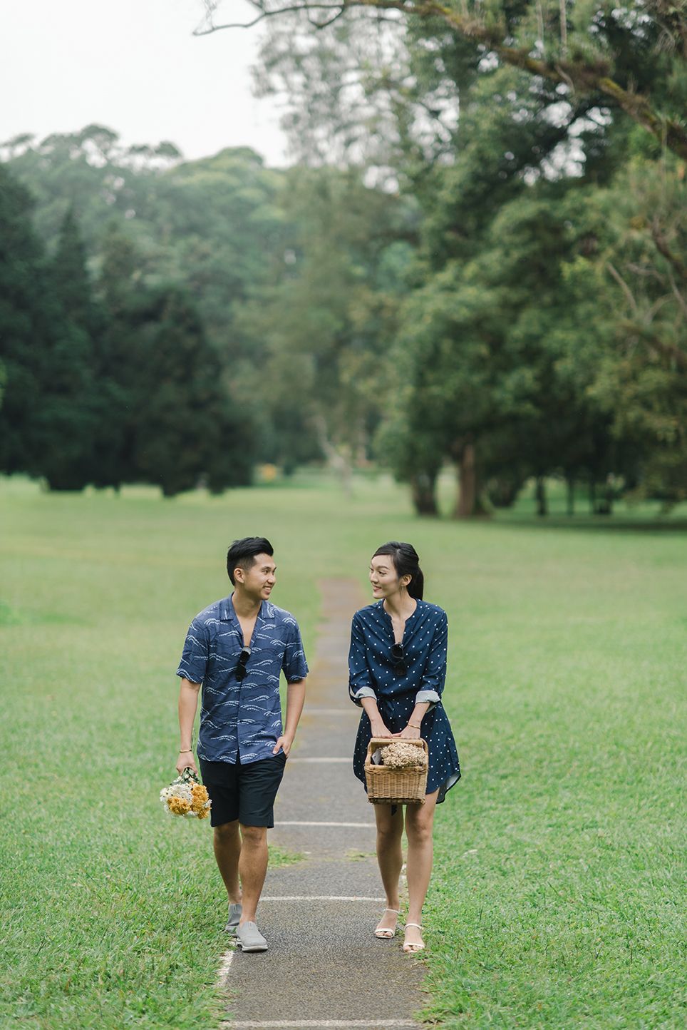 Couple in matching blue outfits walking on a park path, carrying a picnic basket and flowers.