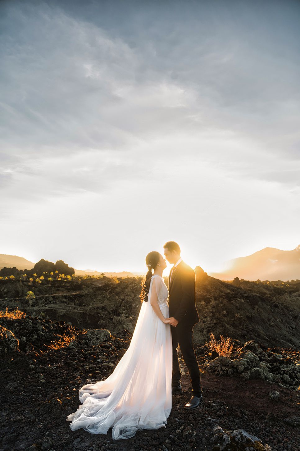 Bride and groom stand together at golden sunset on volcanic landscape, her white gown trailing on dark rocky ground.
