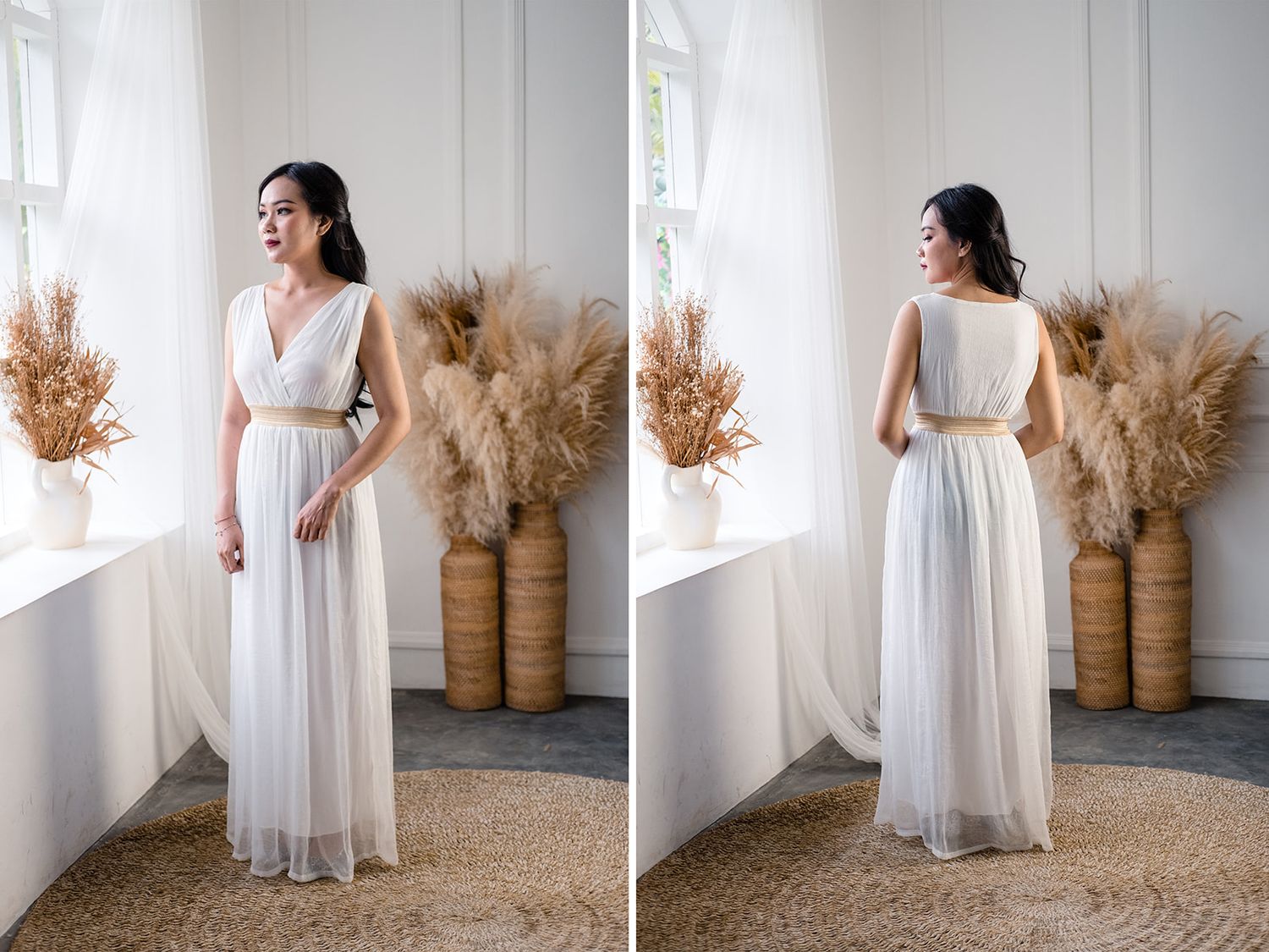 Woman in elegant white chiffon maxi dress with gold belt, posing near window with pampas grass decor.