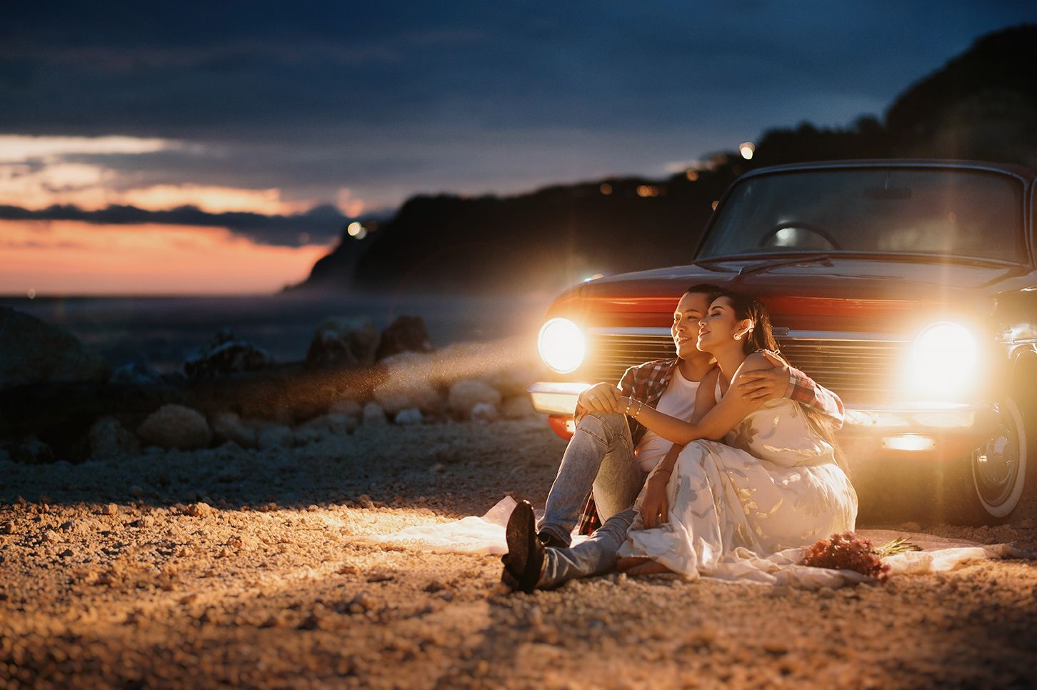Couple sitting together in front of vintage car headlights at sunset on rocky terrain.