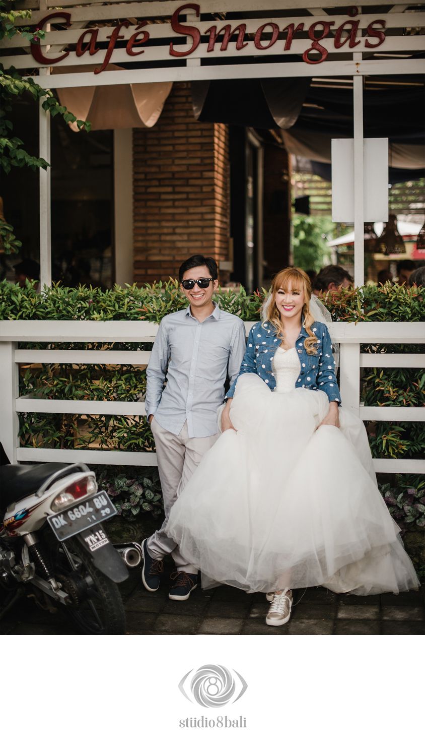 Couple posing in front of Cafe Smorgas, bride in white tulle gown with denim jacket, groom in gray shirt with sunglasses.