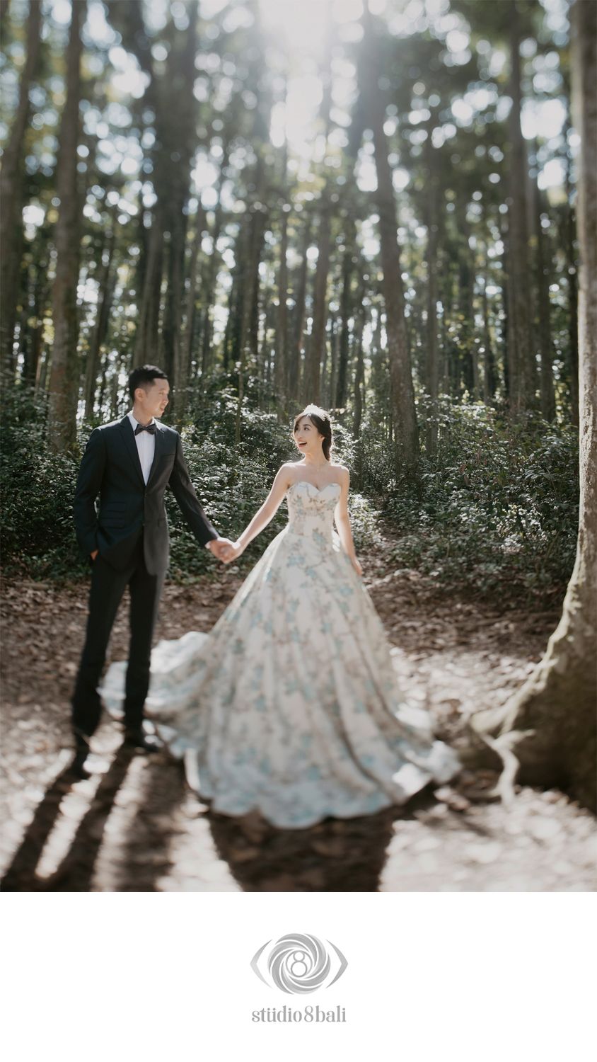 Couple in wedding attire holding hands in a sunlit forest, bride wearing a floral ball gown, groom in a black tuxedo.