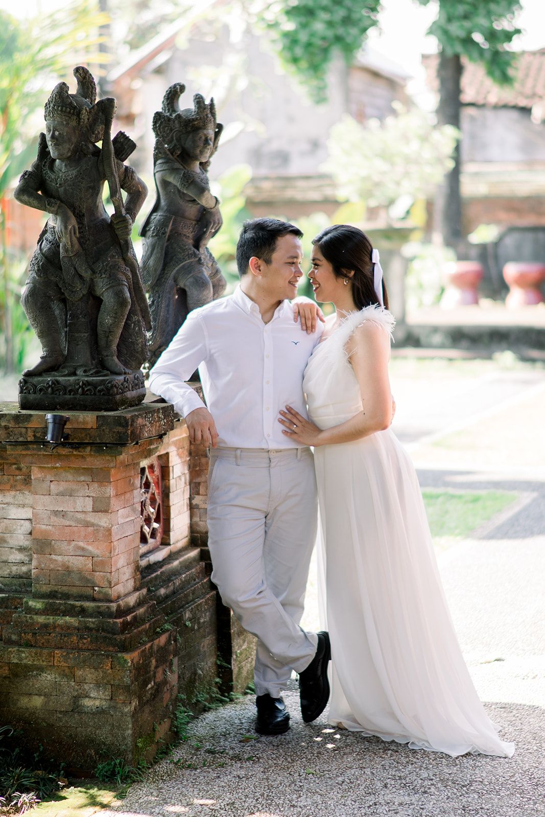 Couple in white wedding attire stands romantically beside ancient Balinese stone statues at a temple.