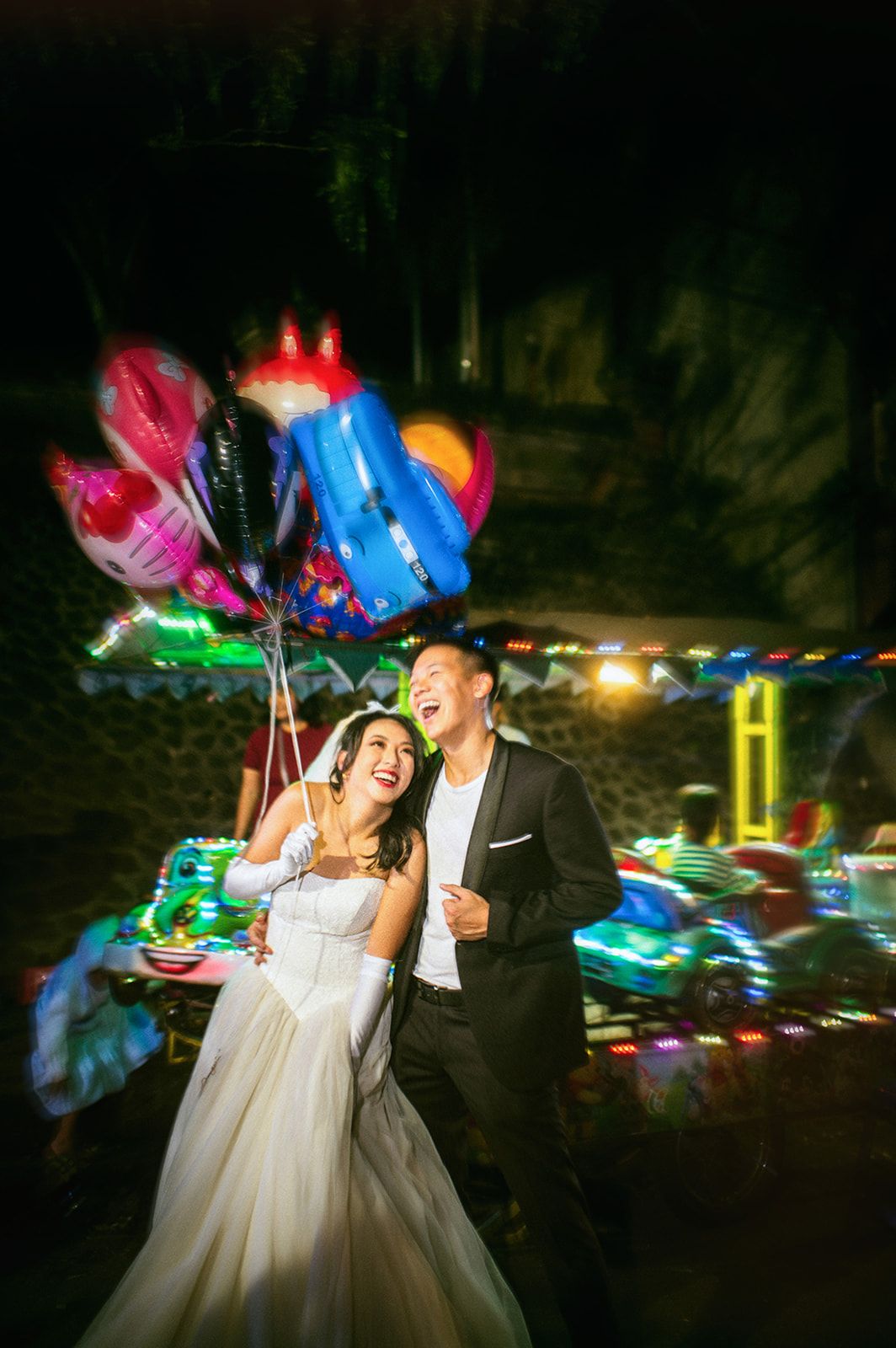 Joyful bride and groom laughing together at night carnival holding colorful balloons in wedding attire.