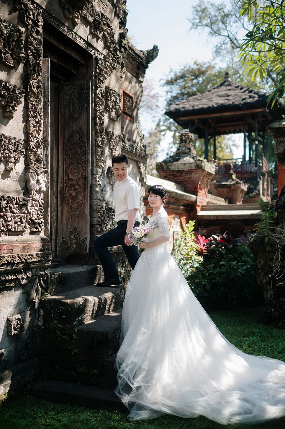 Couple in wedding attire posing at a Balinese temple, bride in white gown with train, groom in white shirt on stone steps.