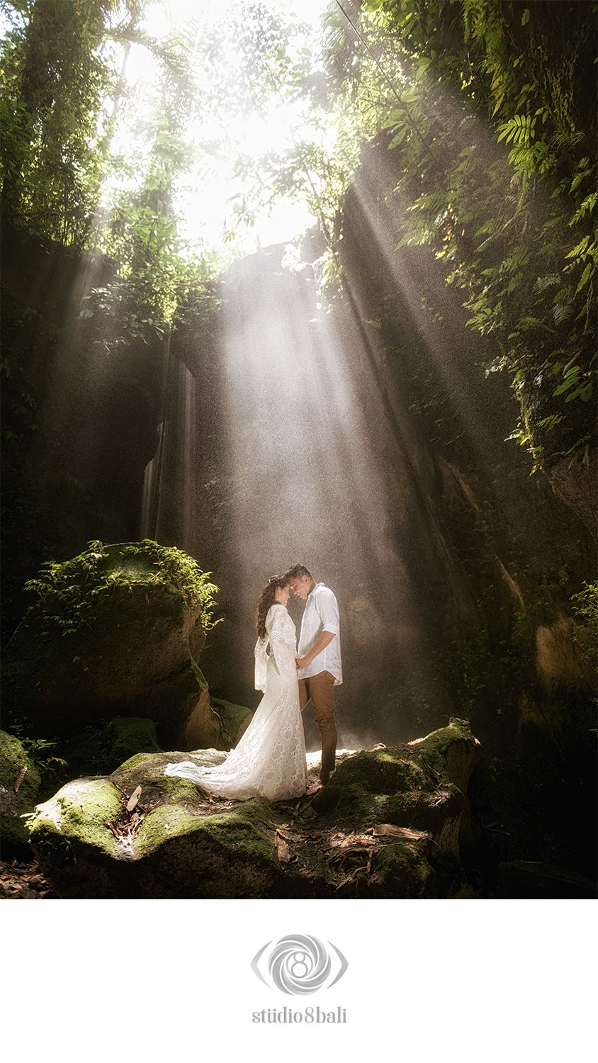 Couple kissing in white wedding attire surrounded by lush greenery and dramatic sunbeams in a magical forest setting.
