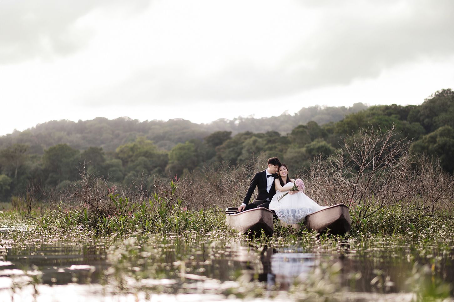 Couple in wedding attire sitting romantically in a canoe surrounded by lush greenery and calm waters.
