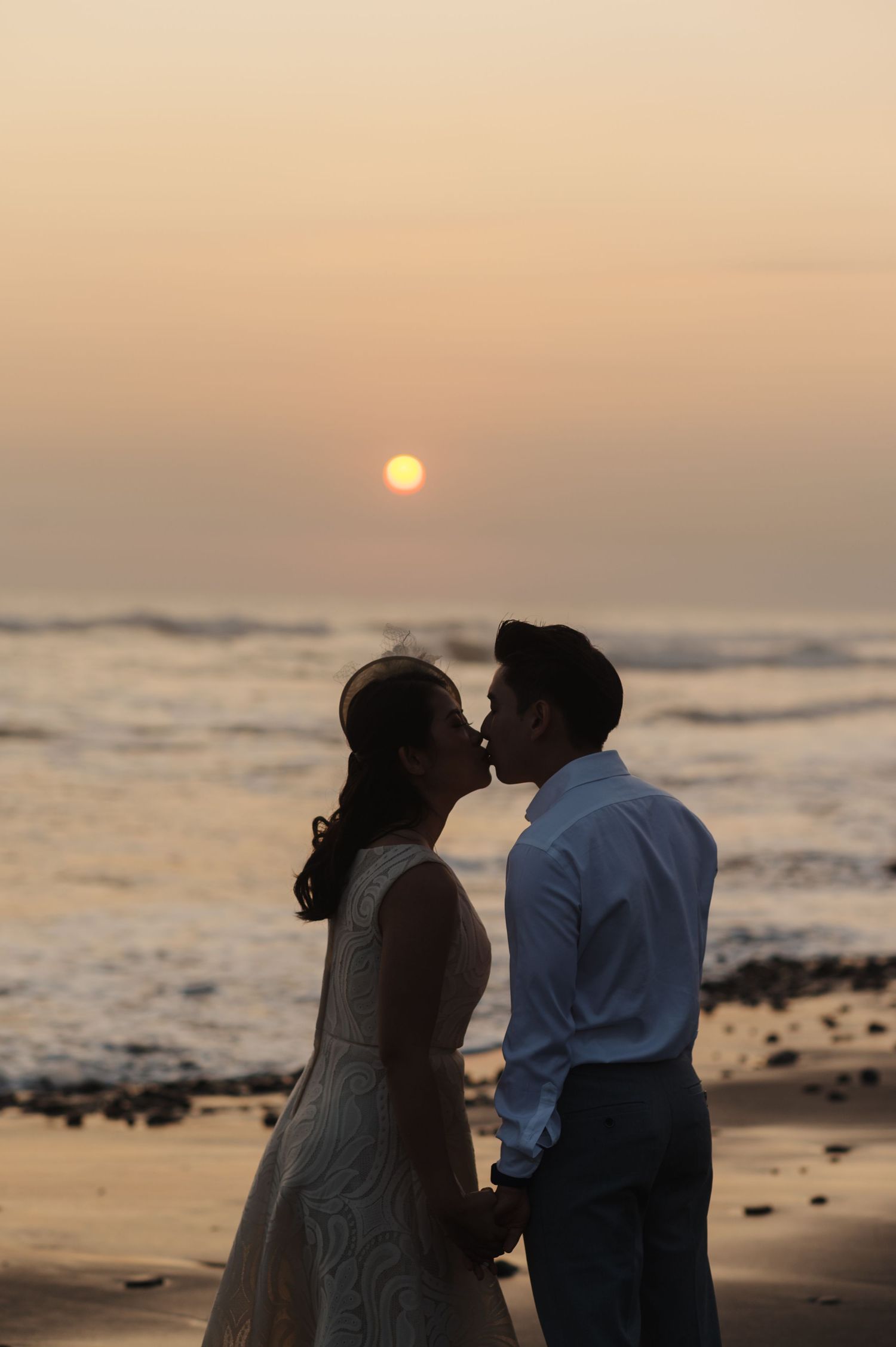Couple sharing a romantic kiss on the beach at sunset, holding hands with golden sky and ocean waves in the background.