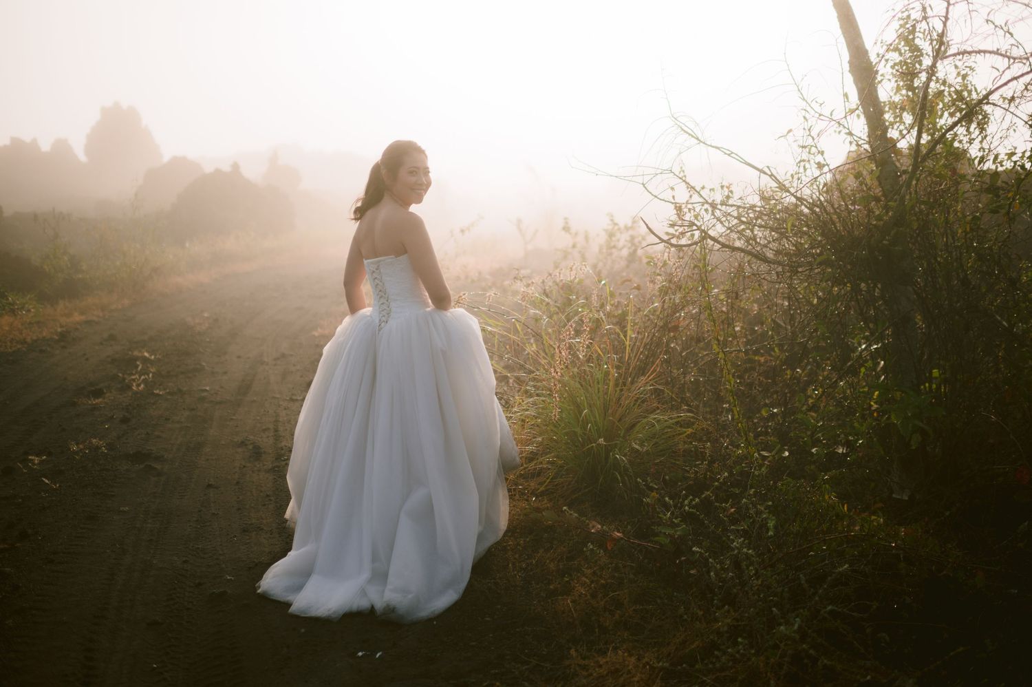 Bride in flowing white tulle gown walks misty rural path at golden hour, soft sunlight illuminating her elegant wedding dress.