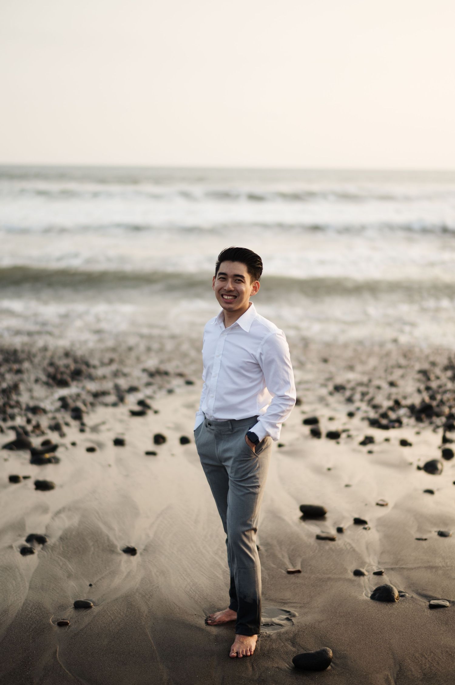 Smiling man in white shirt and gray pants standing barefoot on a sandy beach with ocean waves in the background.