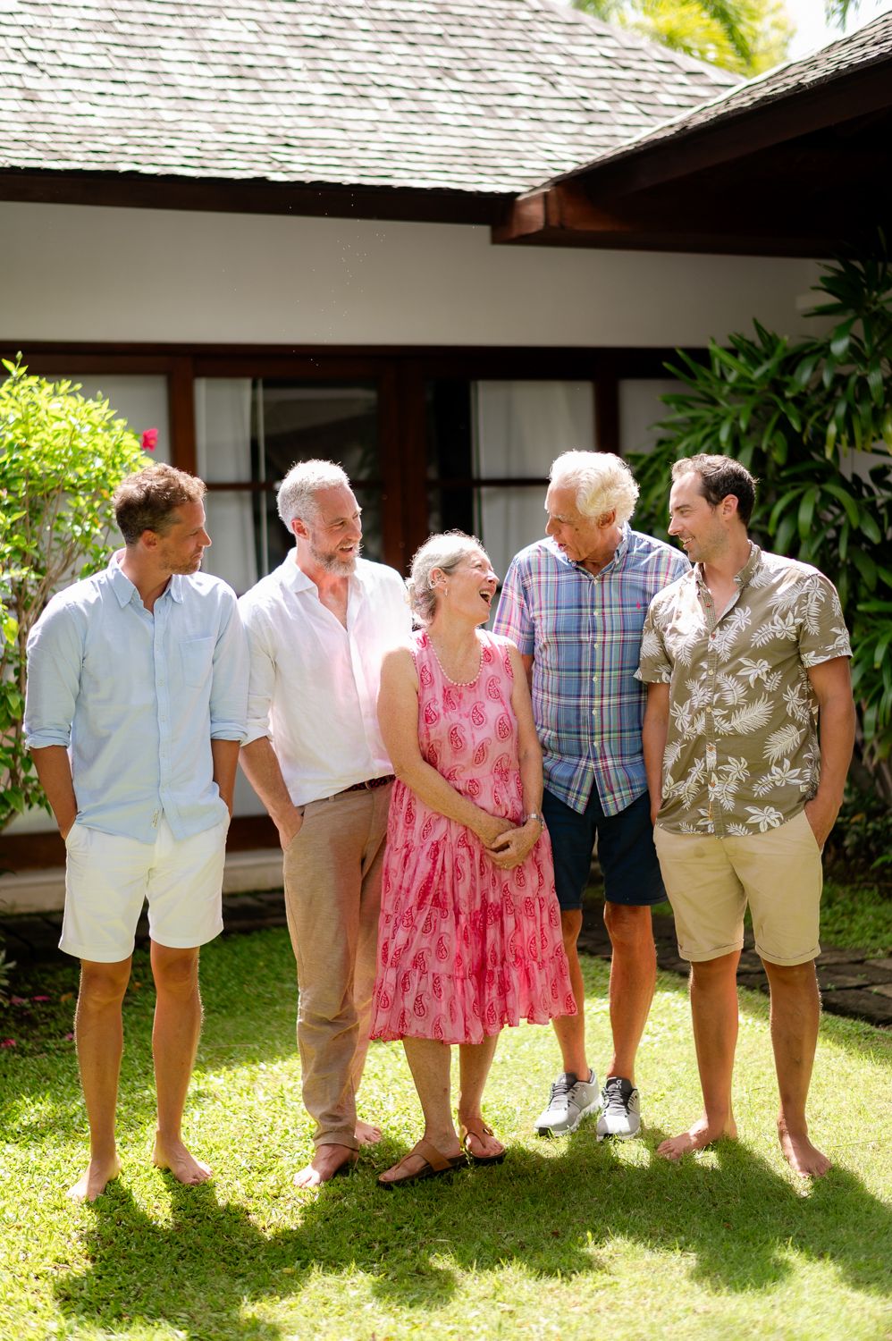 Multi-generational family group smiling together outdoors on green lawn beside tropical vacation villa on sunny day.