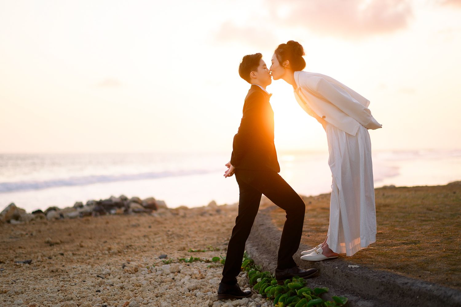 A couple shares a romantic kiss on a sandy beach at sunset, dressed in wedding attire.