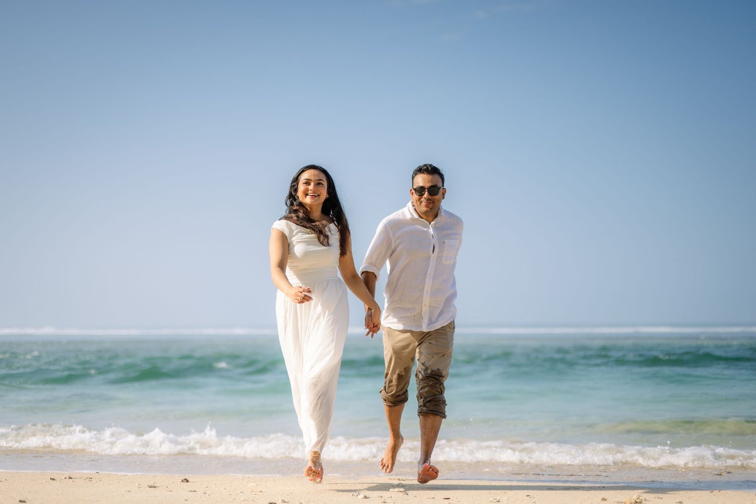 A happy couple holding hands and running barefoot on a sunny beach with turquoise ocean waves in the background.