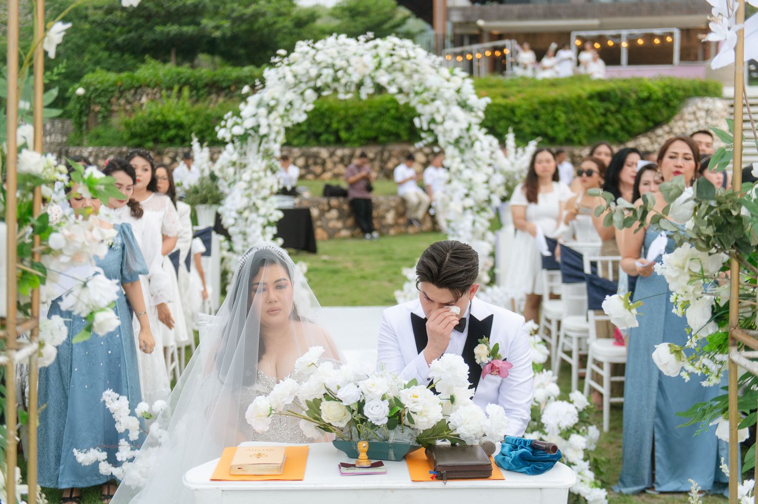 Elegant outdoor wedding ceremony with couple at floral table under white flower arch, bridesmaids in blue dresses lining the aisle.
