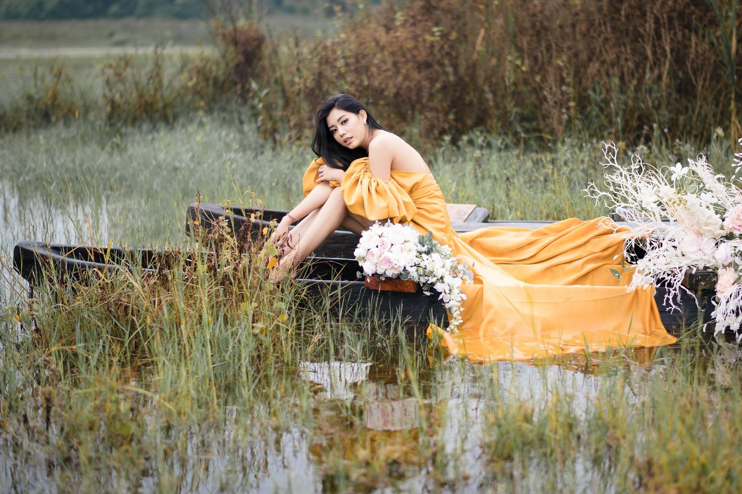 Woman in elegant orange gown reclining in a rustic boat surrounded by wildflowers and tall grass in a serene wetland.