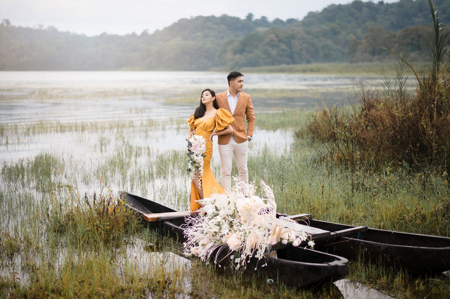 Couple in romantic pre-wedding photoshoot standing on a wooden boat surrounded by flowers on a serene lake.