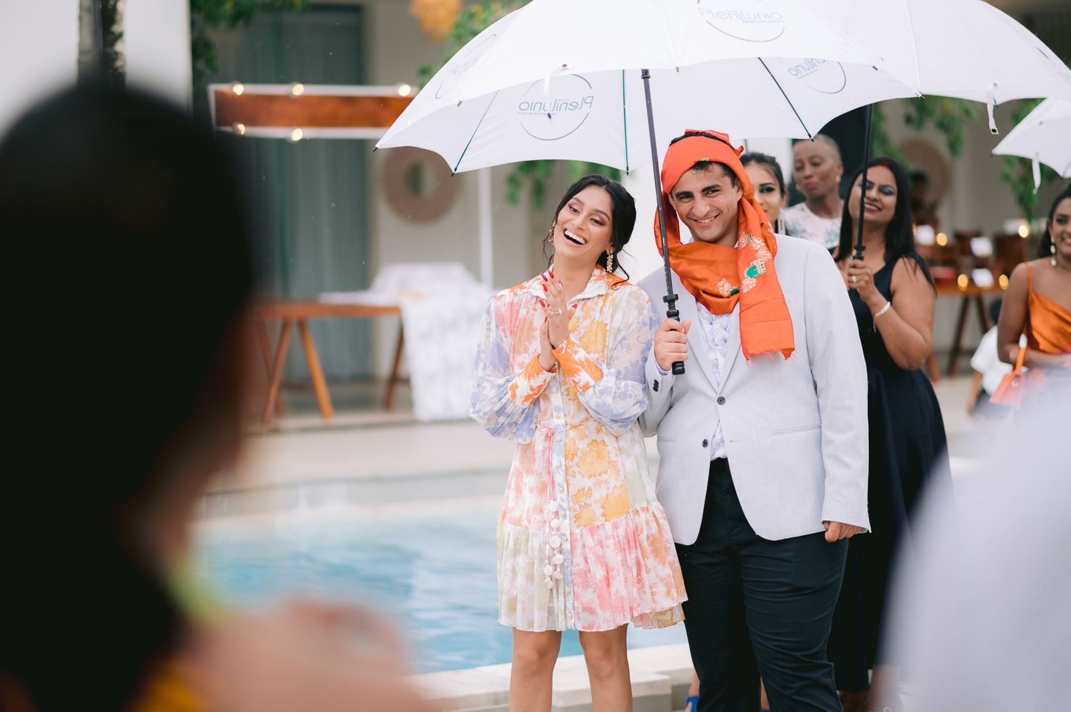 Couple smiles under a white umbrella by the pool, man wearing an orange turban at a wedding.
