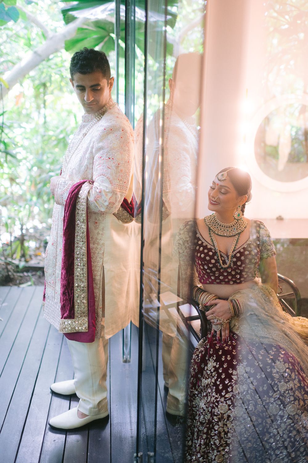 Indian bride in maroon lehenga and groom in white sherwani pose separately in a elegant first-look wedding photo.