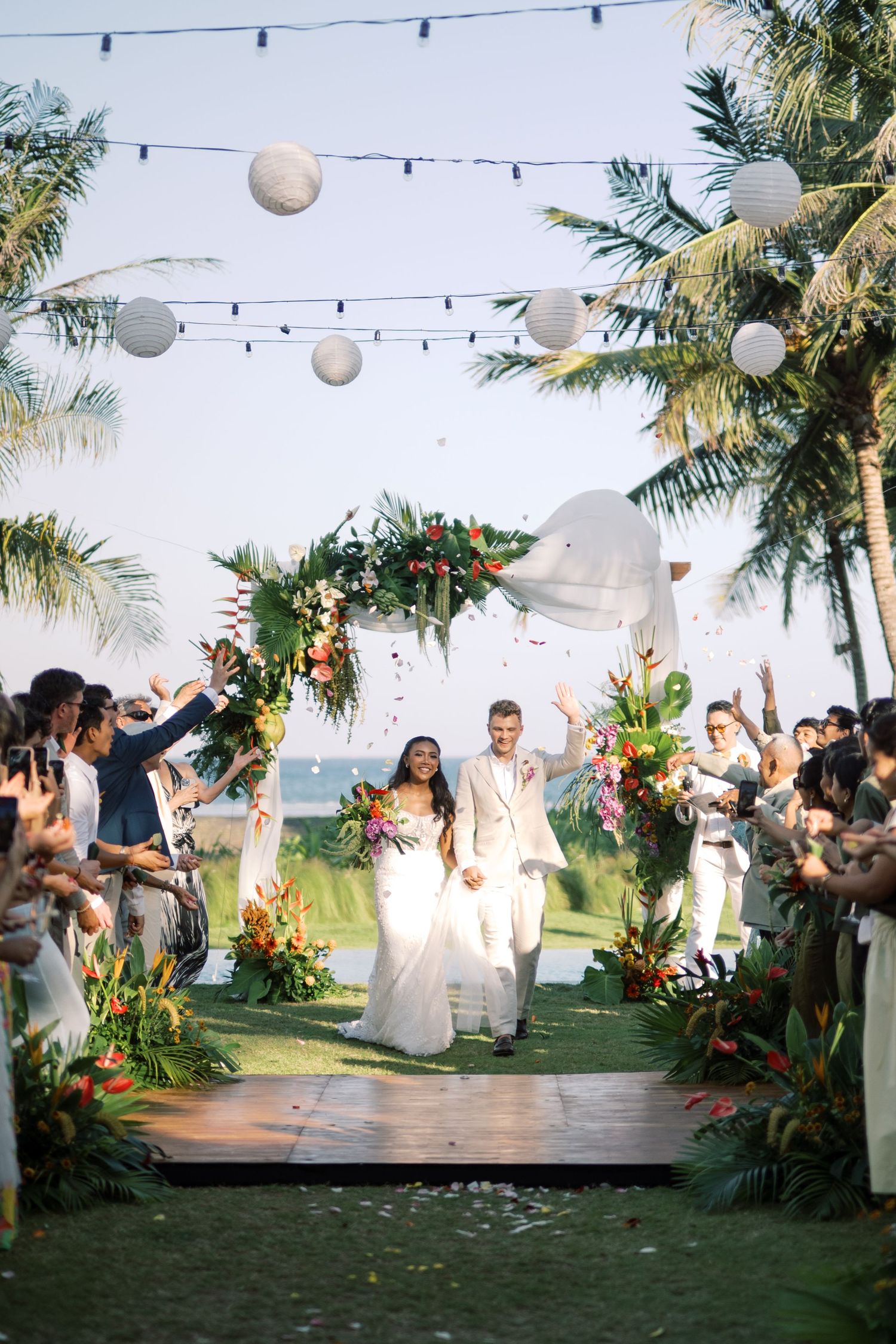 Newlyweds celebrate recessional under floral arch with palm trees and paper lanterns.