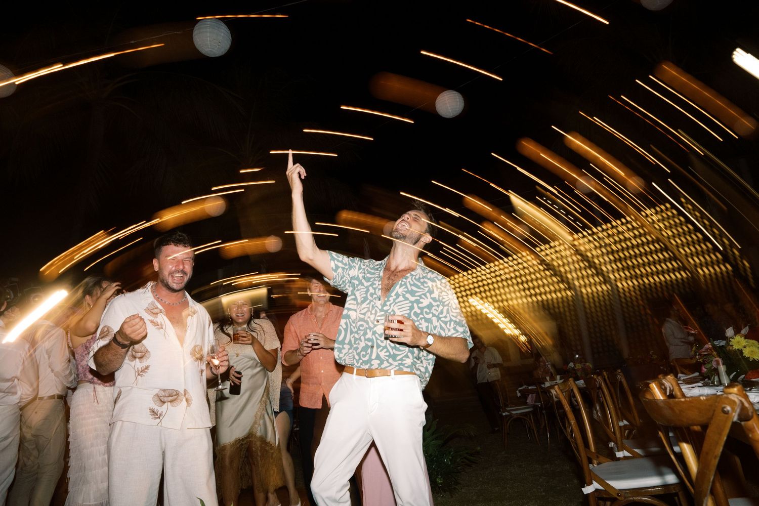 Guests dancing energetically under light trails at lively outdoor wedding reception at night.