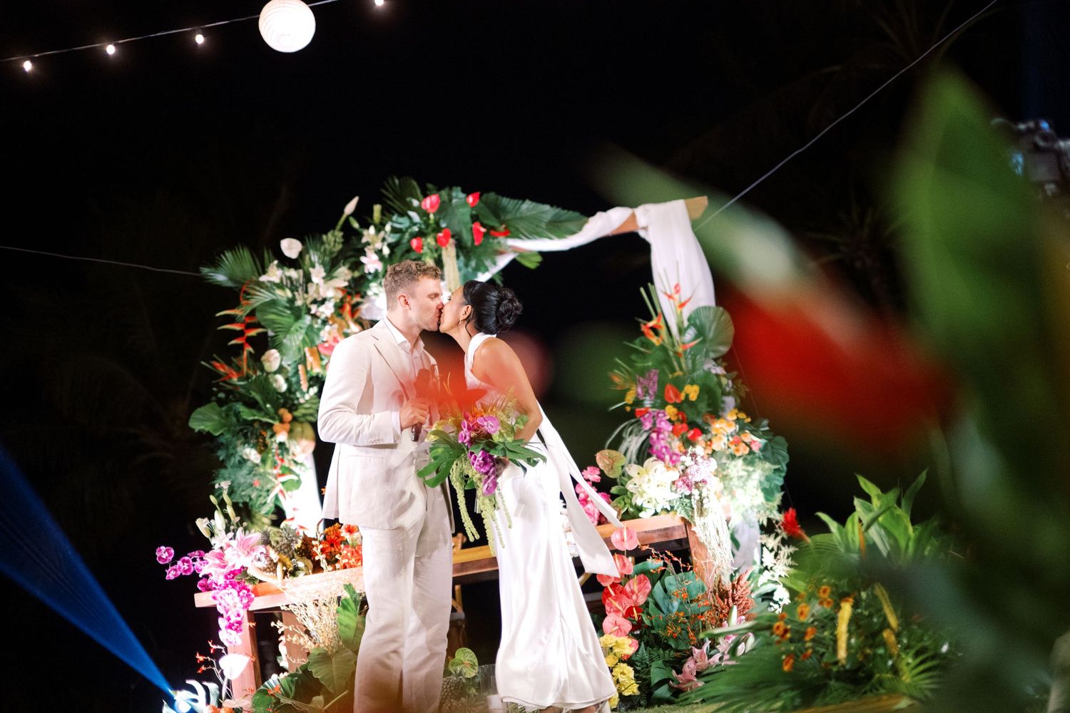 Couple kisses under vibrant tropical floral arch at outdoor night wedding ceremony.