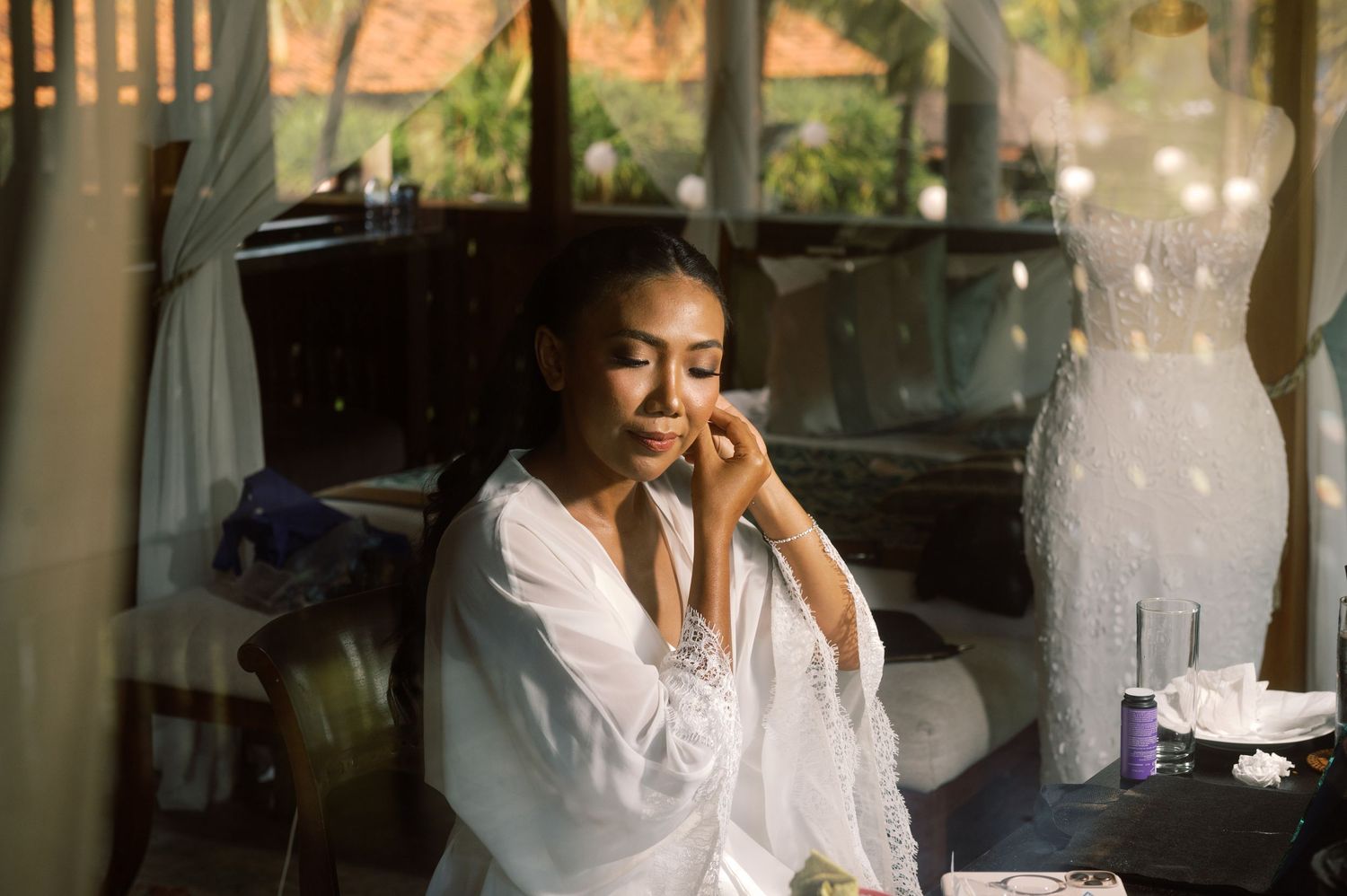 Bride in white robe applying makeup during wedding preparations in a tropical villa.