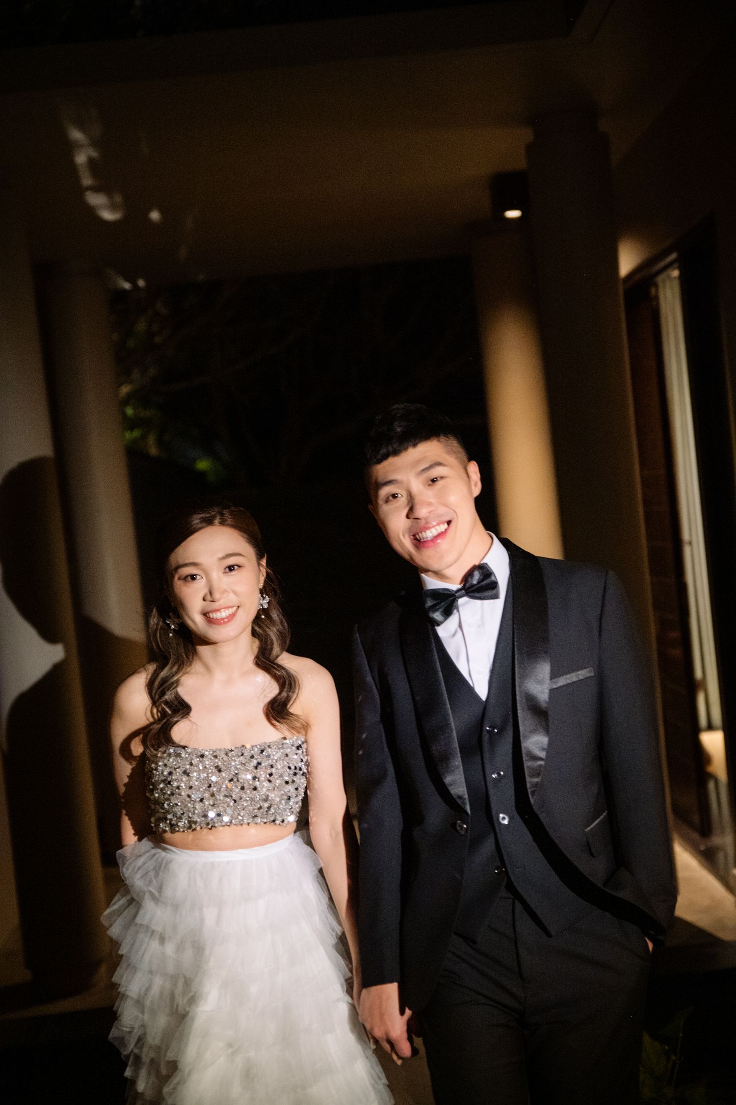 A smiling couple holds hands in formal evening attire, she in a sparkly beaded top and tulle skirt, he in a black tuxedo with bow tie.