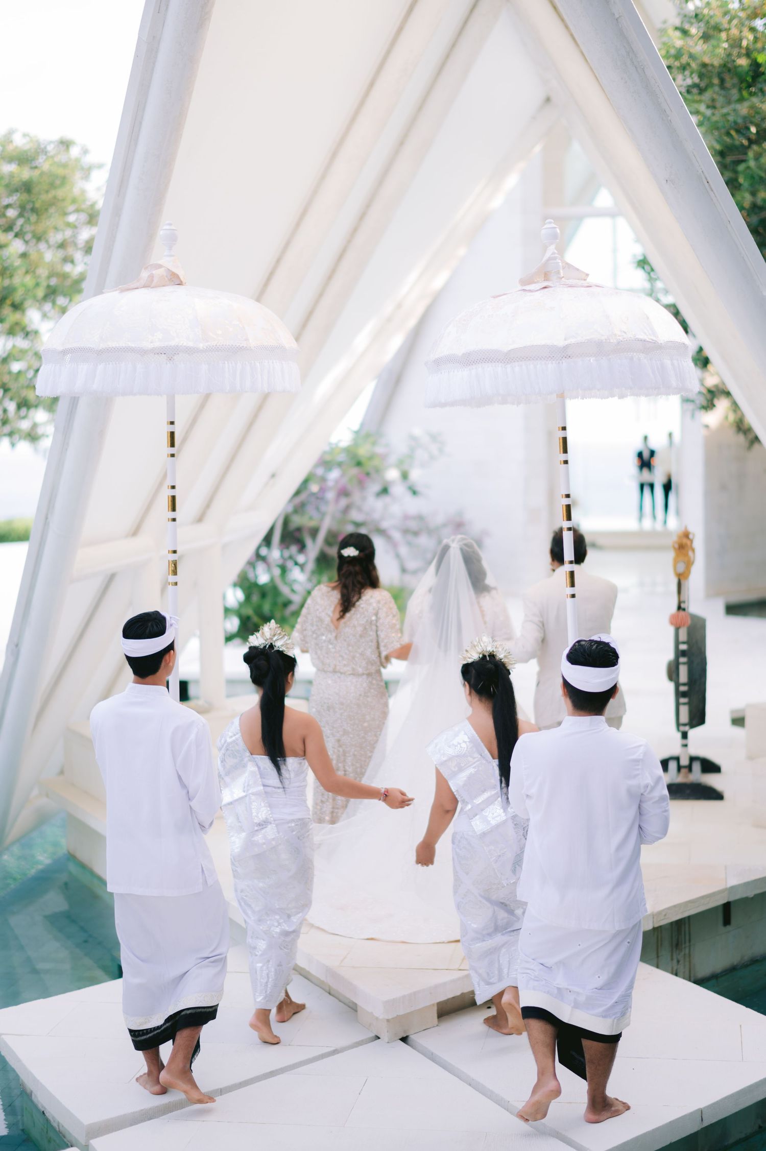 Balinese wedding ceremony with guests in white traditional attire approaching a white open-air chapel with decorative umbrellas.