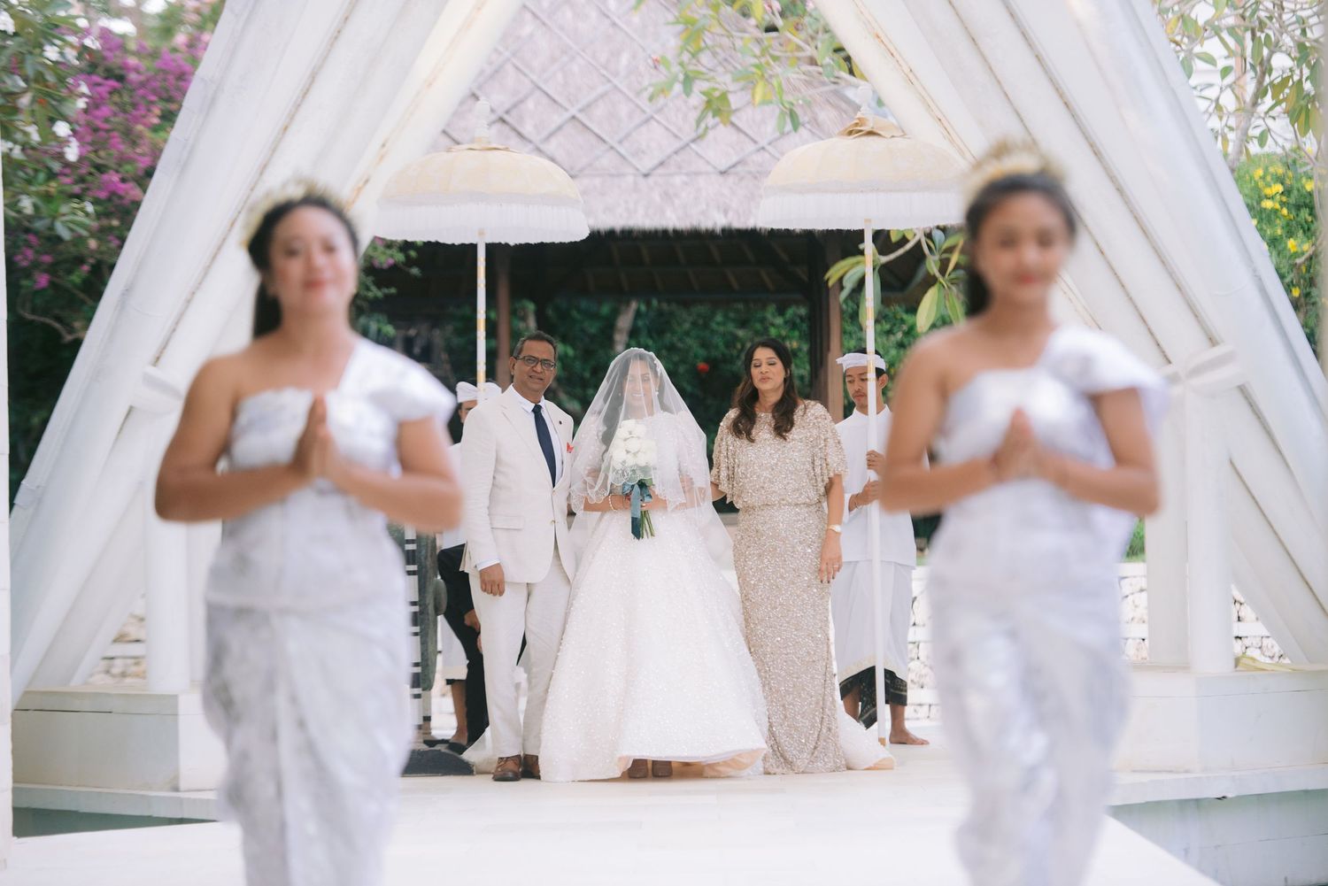 Elegant outdoor wedding ceremony with bride in white gown, veil, and bridesmaids in silver lace dresses under a white canopy.
