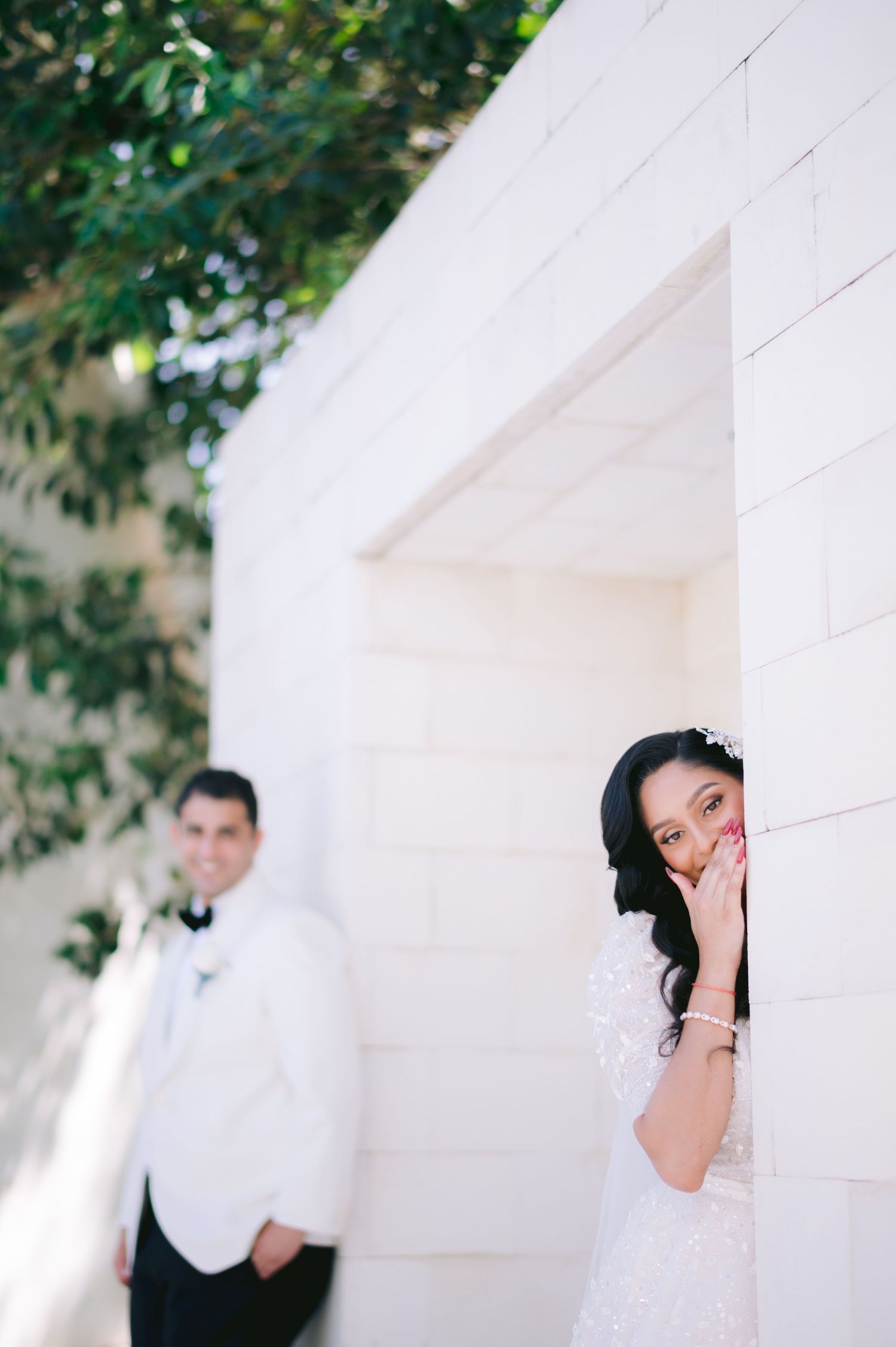 Bride playfully peeks around a white brick wall while groom stands blurred in background during wedding photo session.