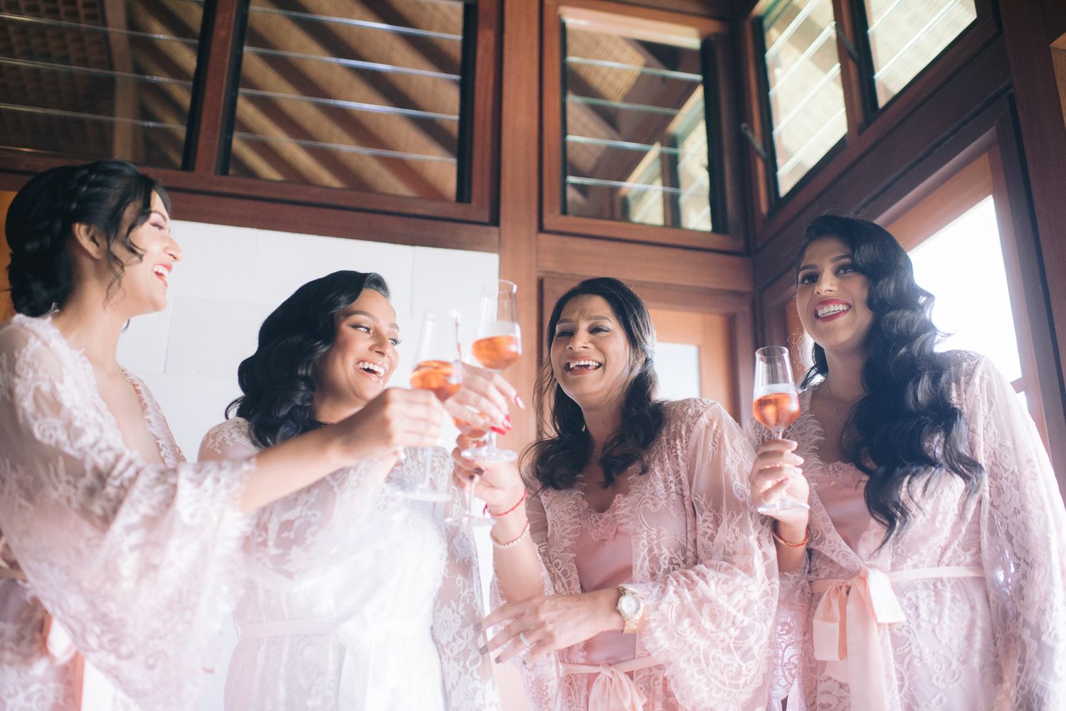 Four bridesmaids in pink lace robes toasting with champagne glasses, laughing together during wedding preparations.