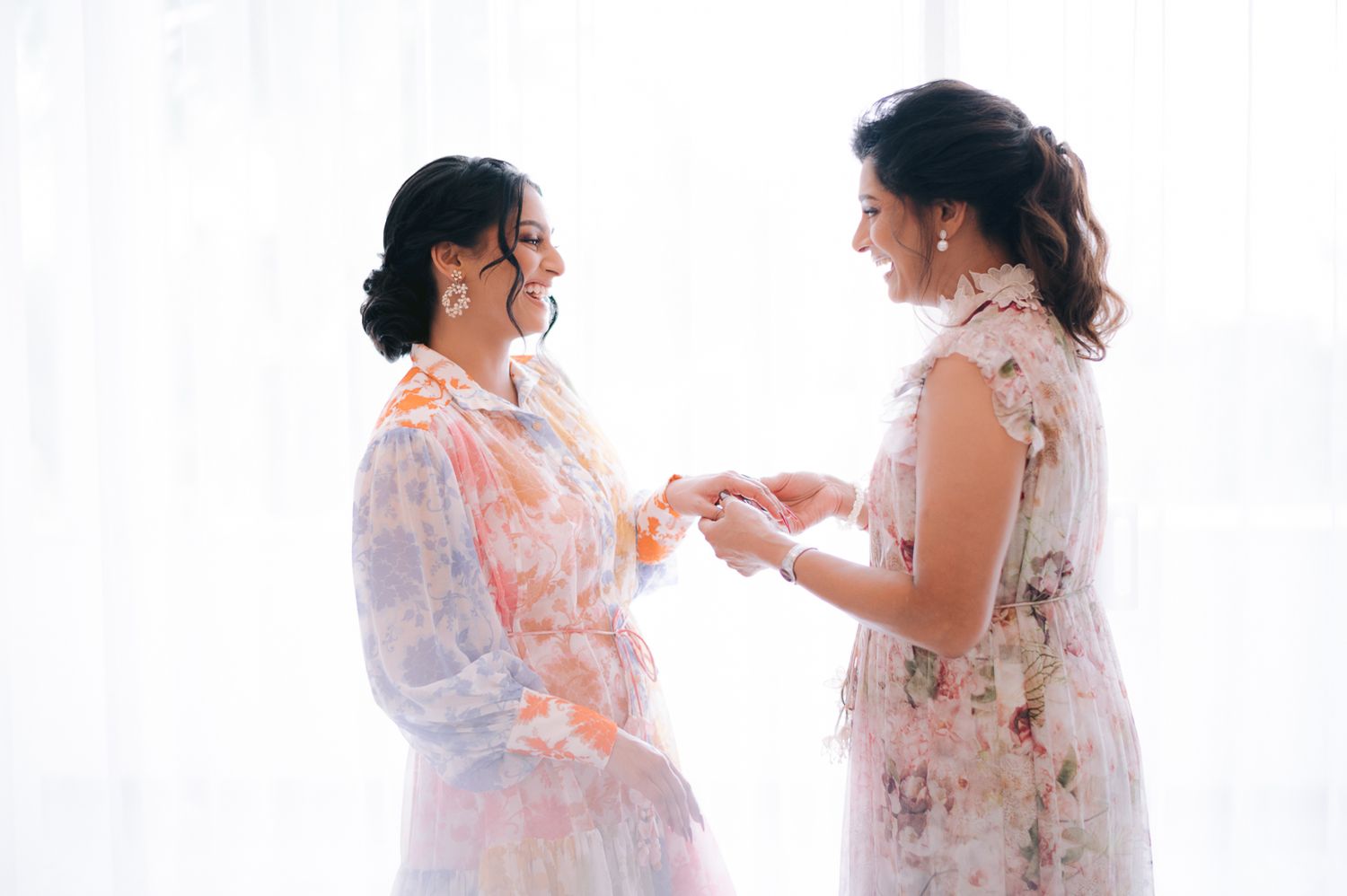 Two women in floral robes share a joyful moment exchanging jewelry during bridal wedding preparations.