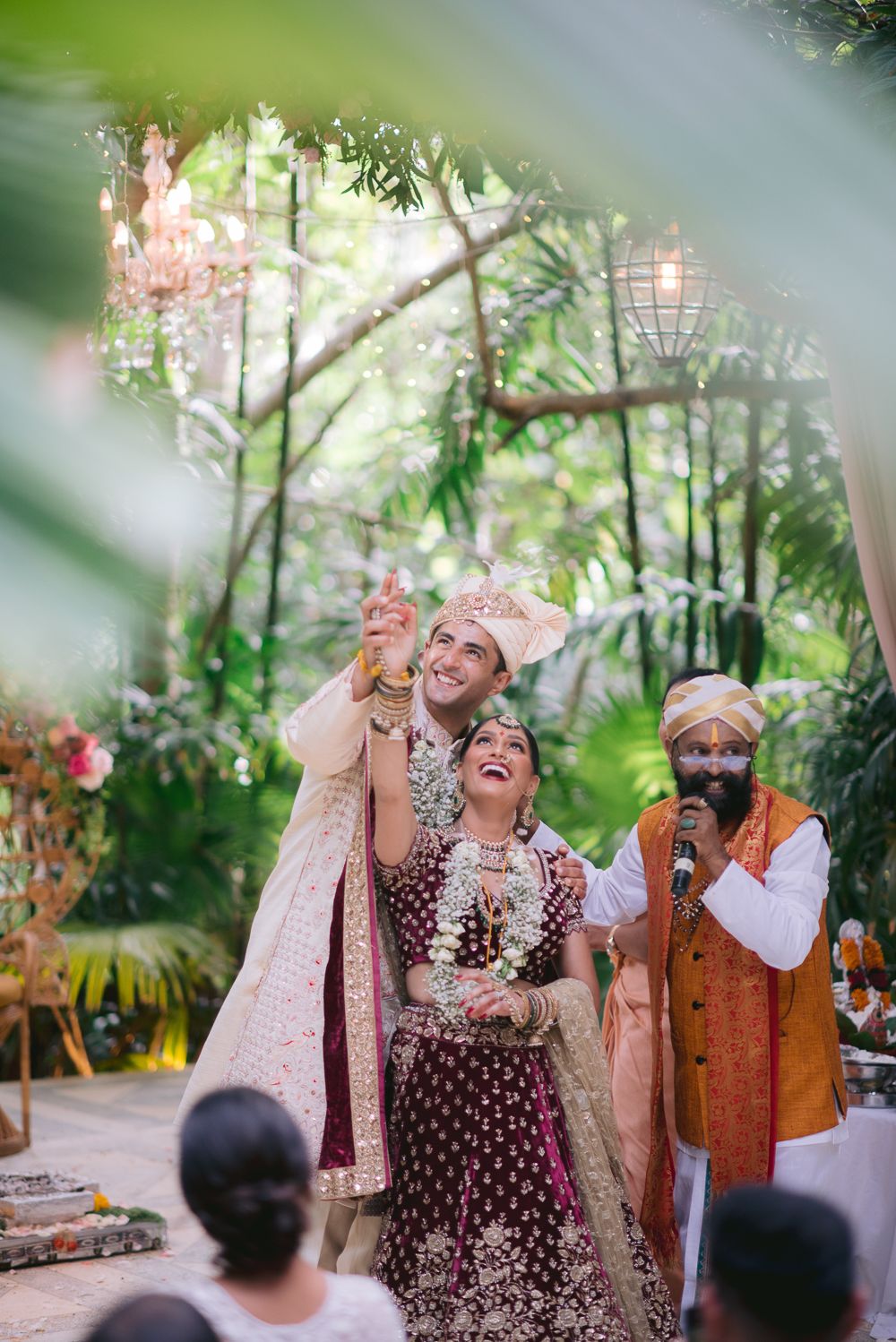 Indian bride and groom laughing joyfully during outdoor wedding ceremony surrounded by lush greenery and floral decor.