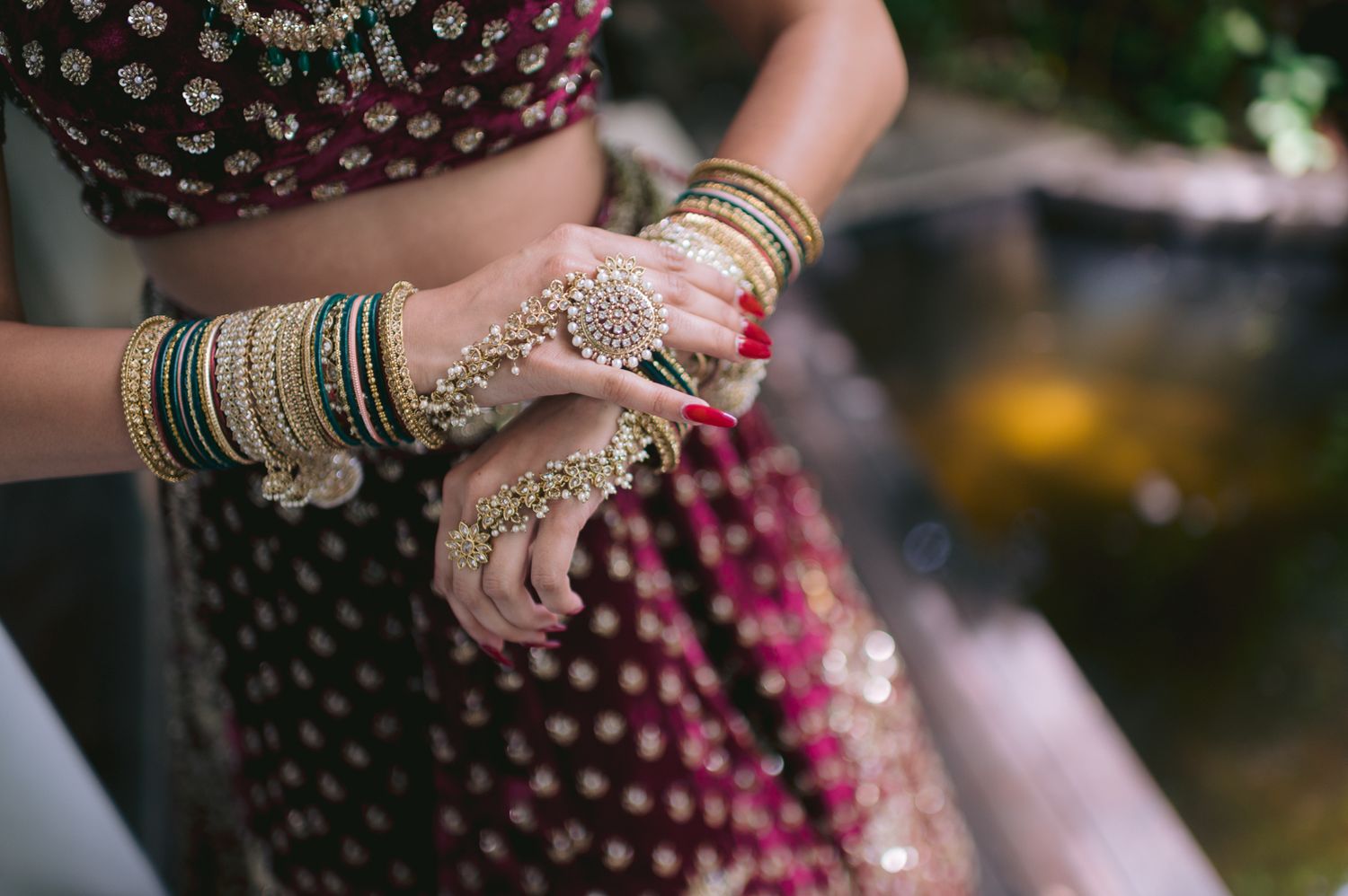 Close-up of Indian bride's hands adorned with gold bangles, mehndi jewelry, and red nail polish on a lehenga.