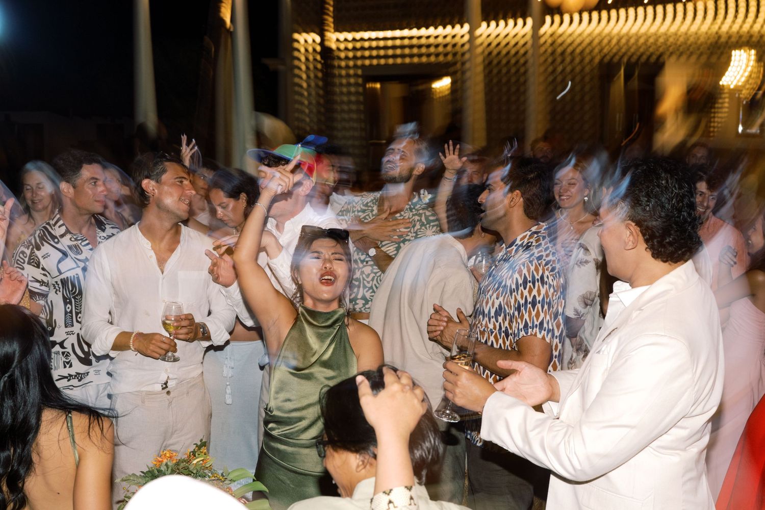Wedding guests dance and celebrate energetically at lively tropical outdoor wedding reception party at night.