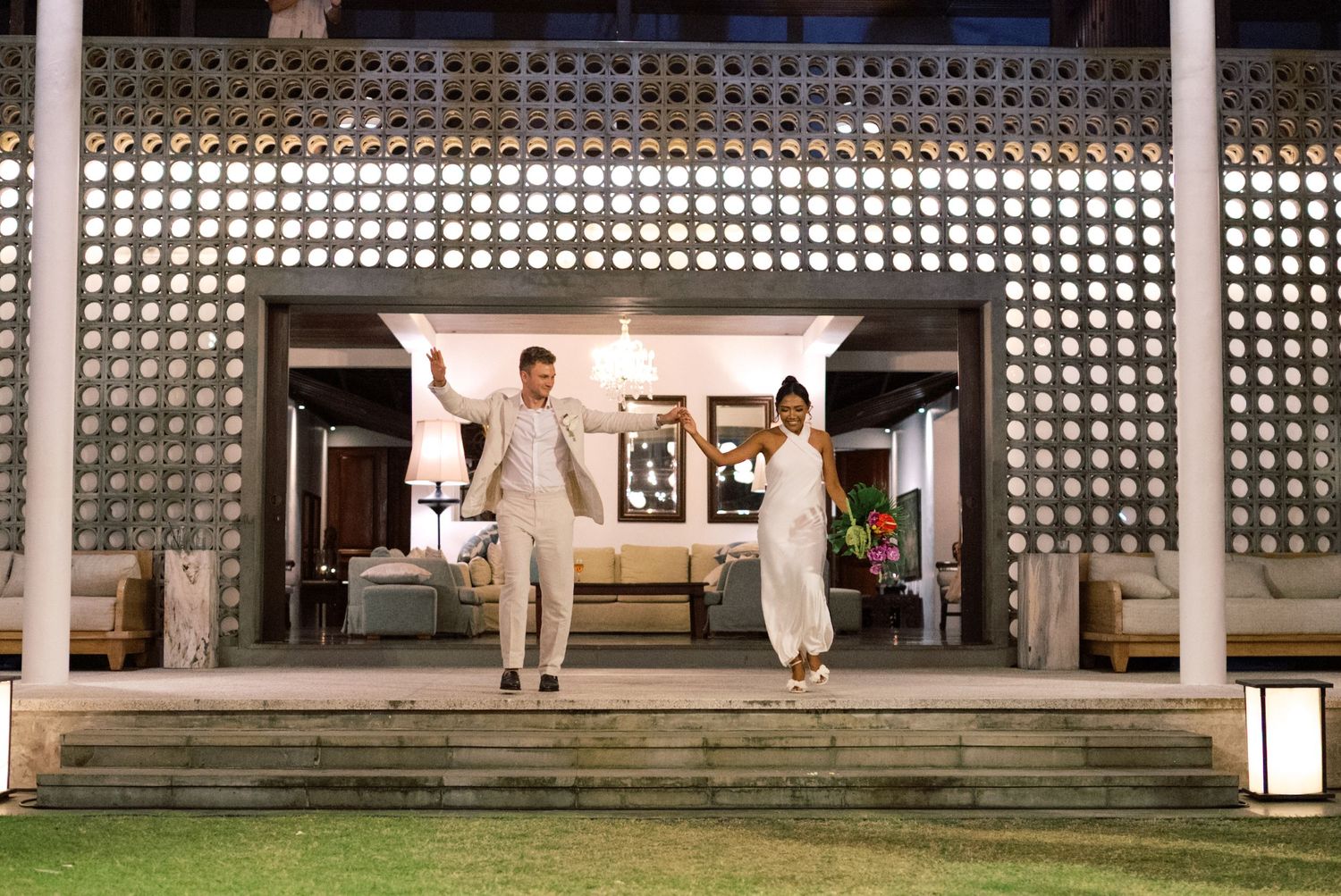 Newlywed couple joyfully descending steps of a lit villa at their tropical evening reception.