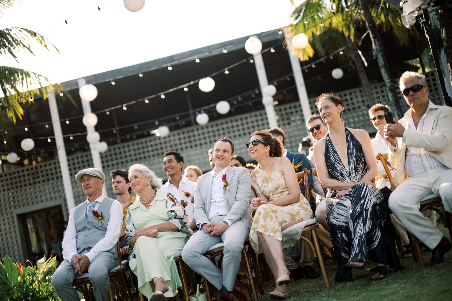 Guests seated outdoors at a tropical wedding ceremony with string lights and white paper lantern decorations.