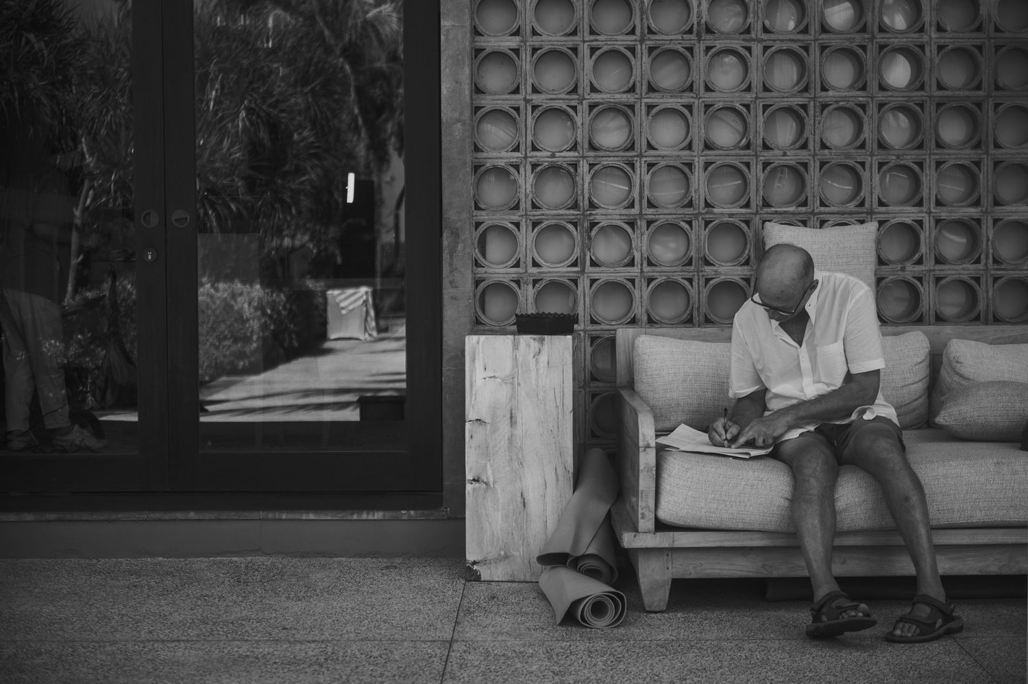Black and white photo of a bald man sitting on an outdoor sofa, reading near a decorative concrete wall.